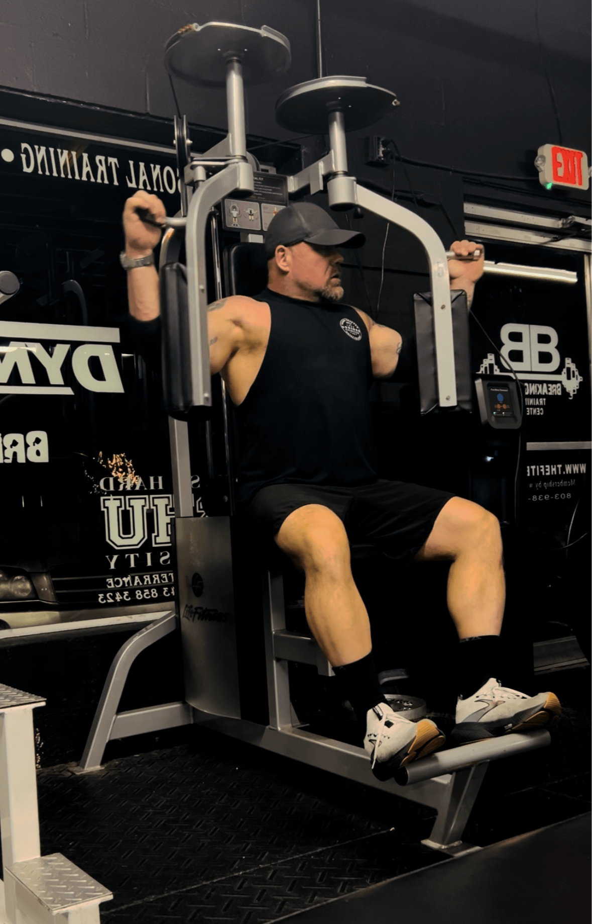 Person sitting on gym equipment, wearing black shirt and shorts, in a gym with black walls and motivational posters.