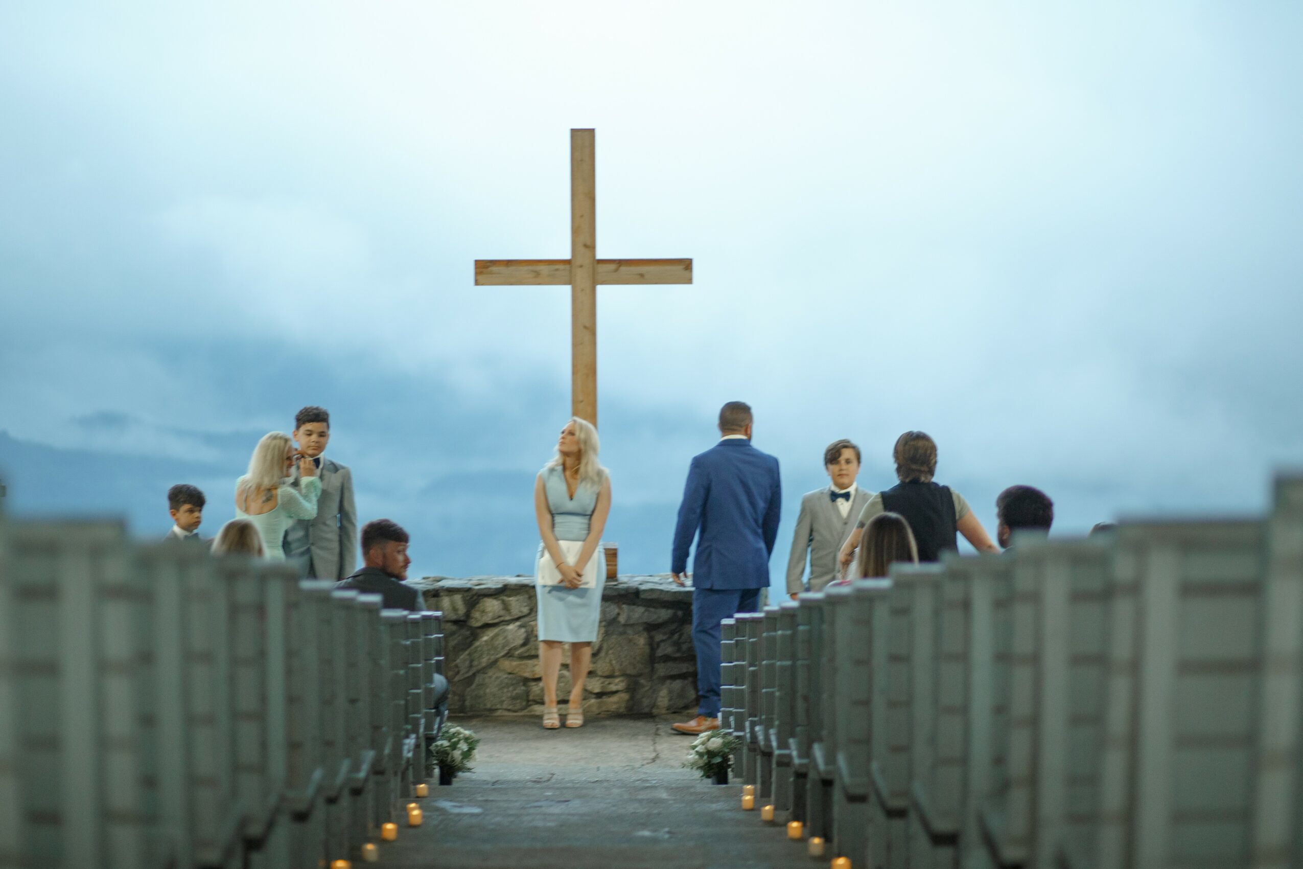 People gathered outdoors near a large cross, with rows of chairs and cloudy sky in the background.