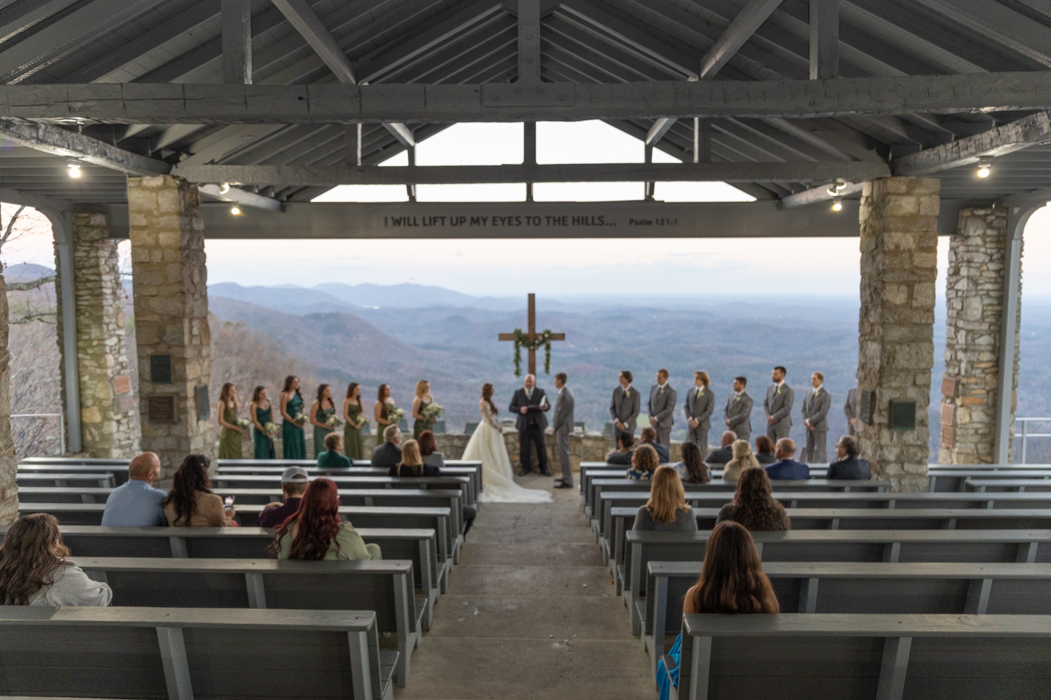 People gather in a chapel with a cross, overlooking mountains and a valley, during daytime. Some are seated, others stand.