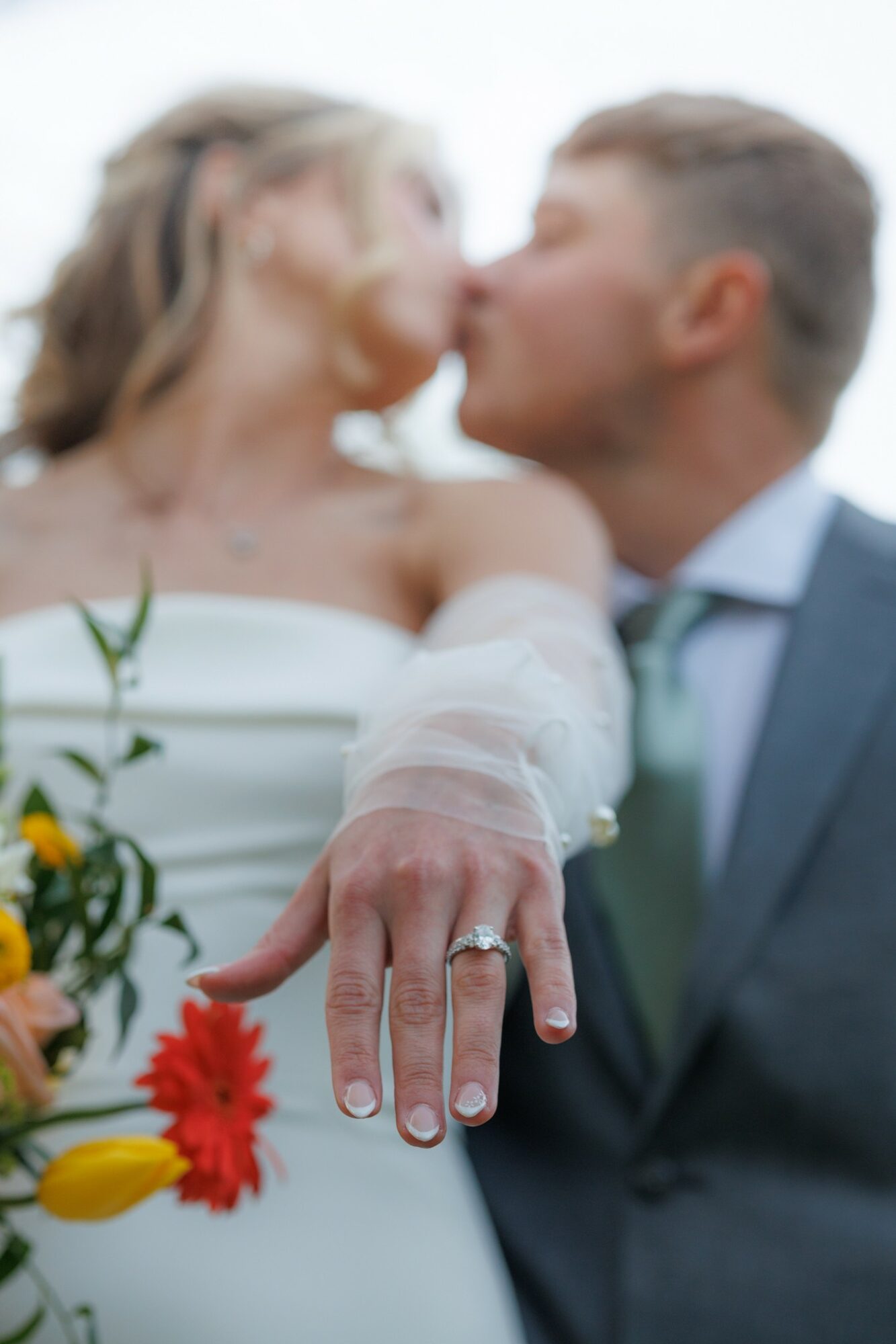 Bride and groom kiss, bride's hand with wedding ring extended forward, holding a bouquet of flowers.