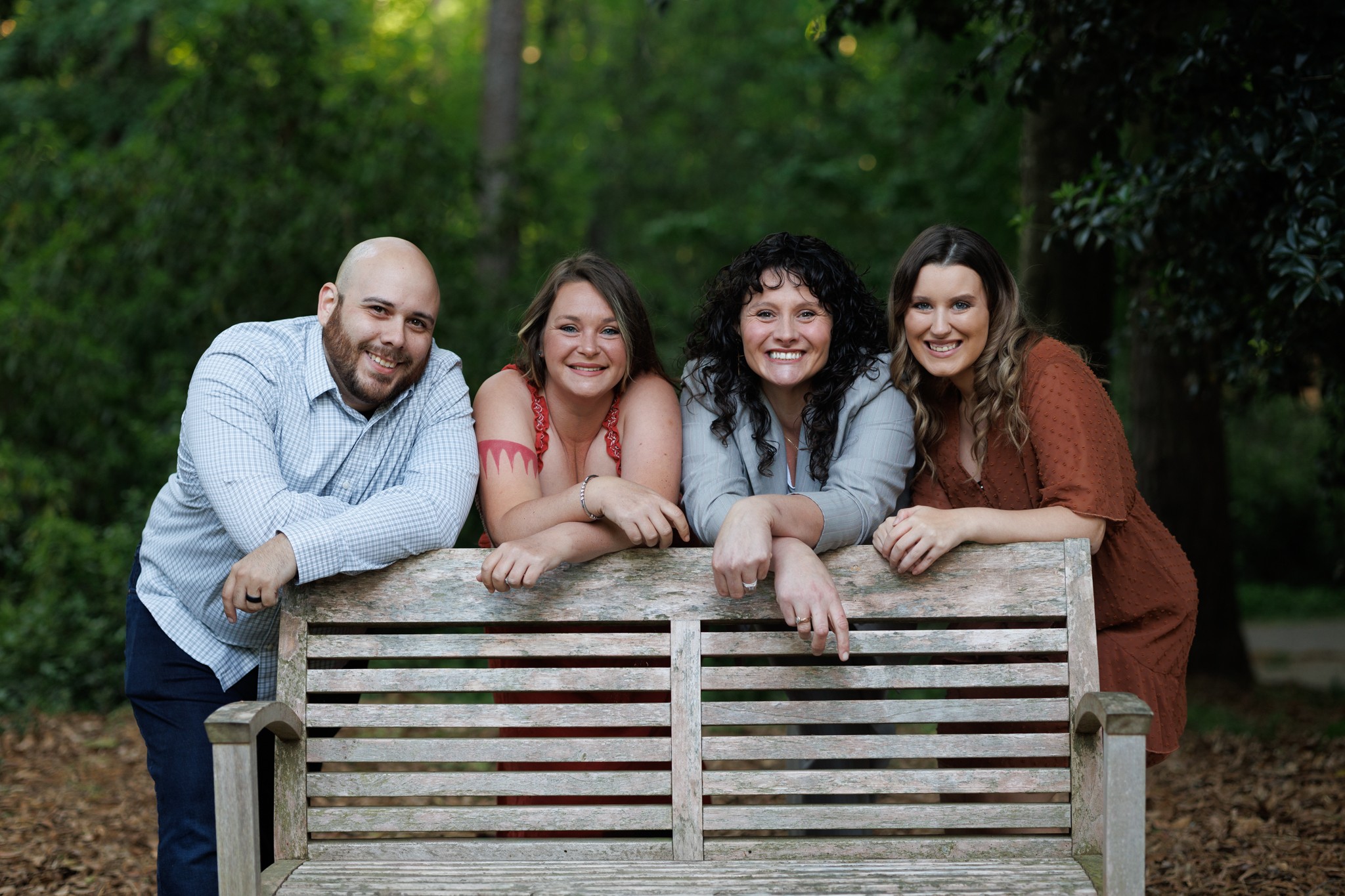 Four people leaning on a wooden bench outdoors, smiling, with trees in the background.
