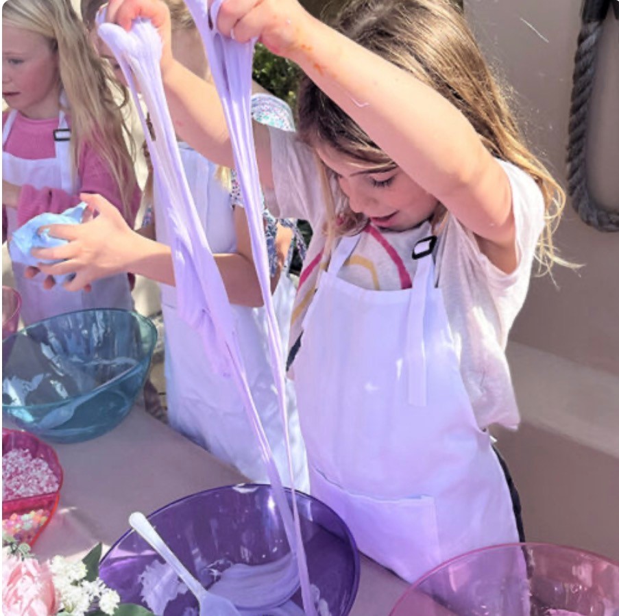 Child with long hair in a white apron pulls stretchy purple slime with other children nearby.