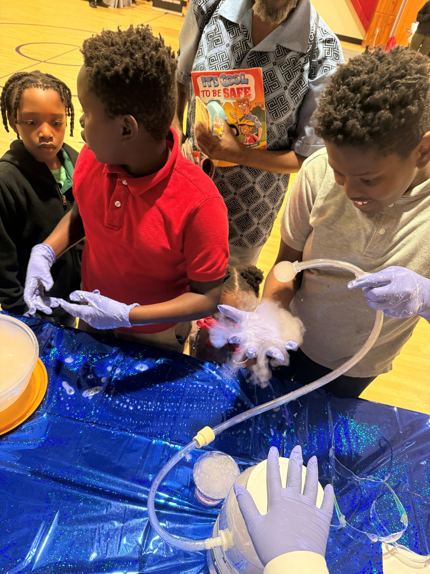 Children and adult gather around a table with a blue cloth, observing a small animal in a container with fog.