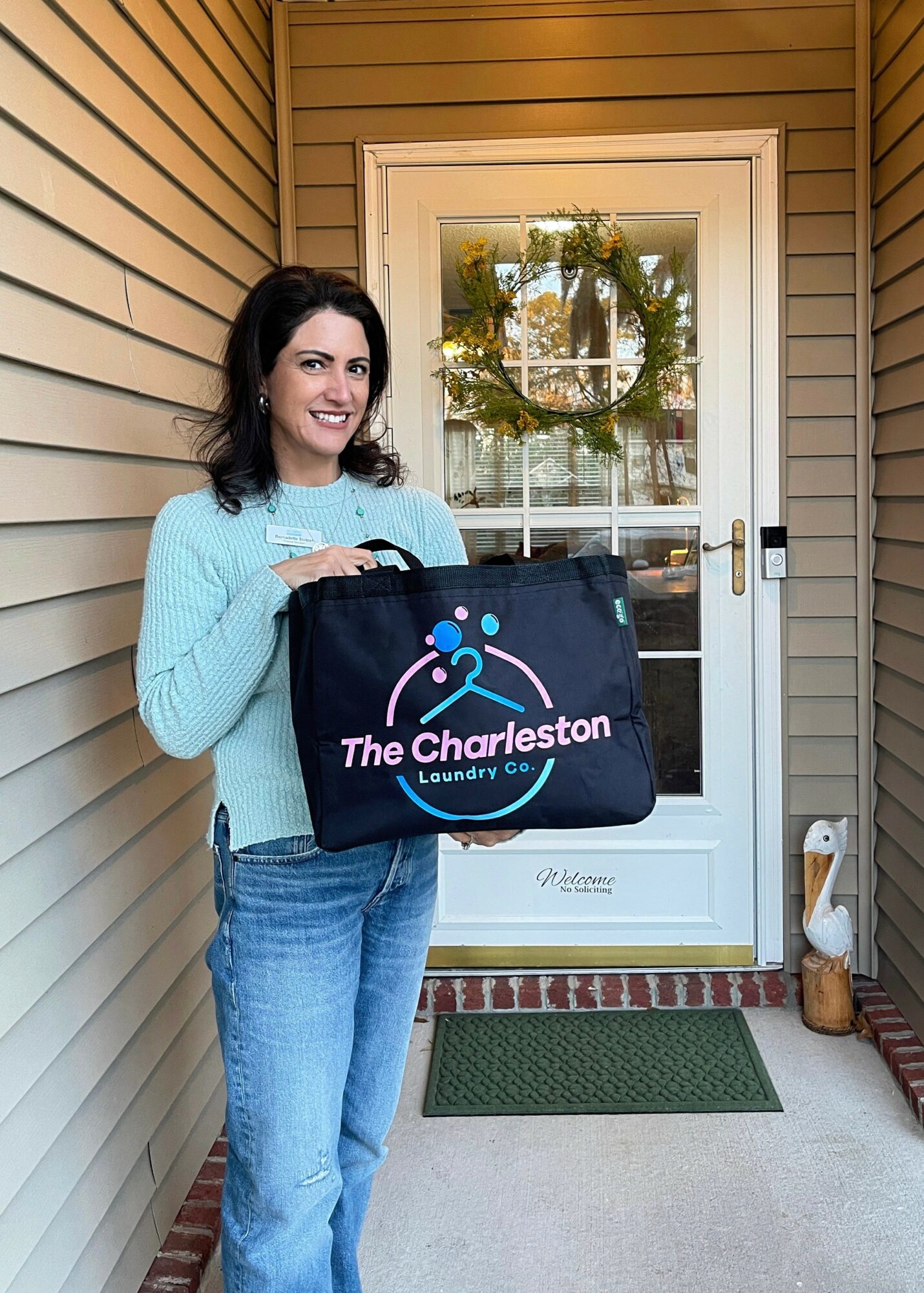 Woman holding a bag with 'The Charleston' logo standing outside a door with a wreath, next to a duck statue.