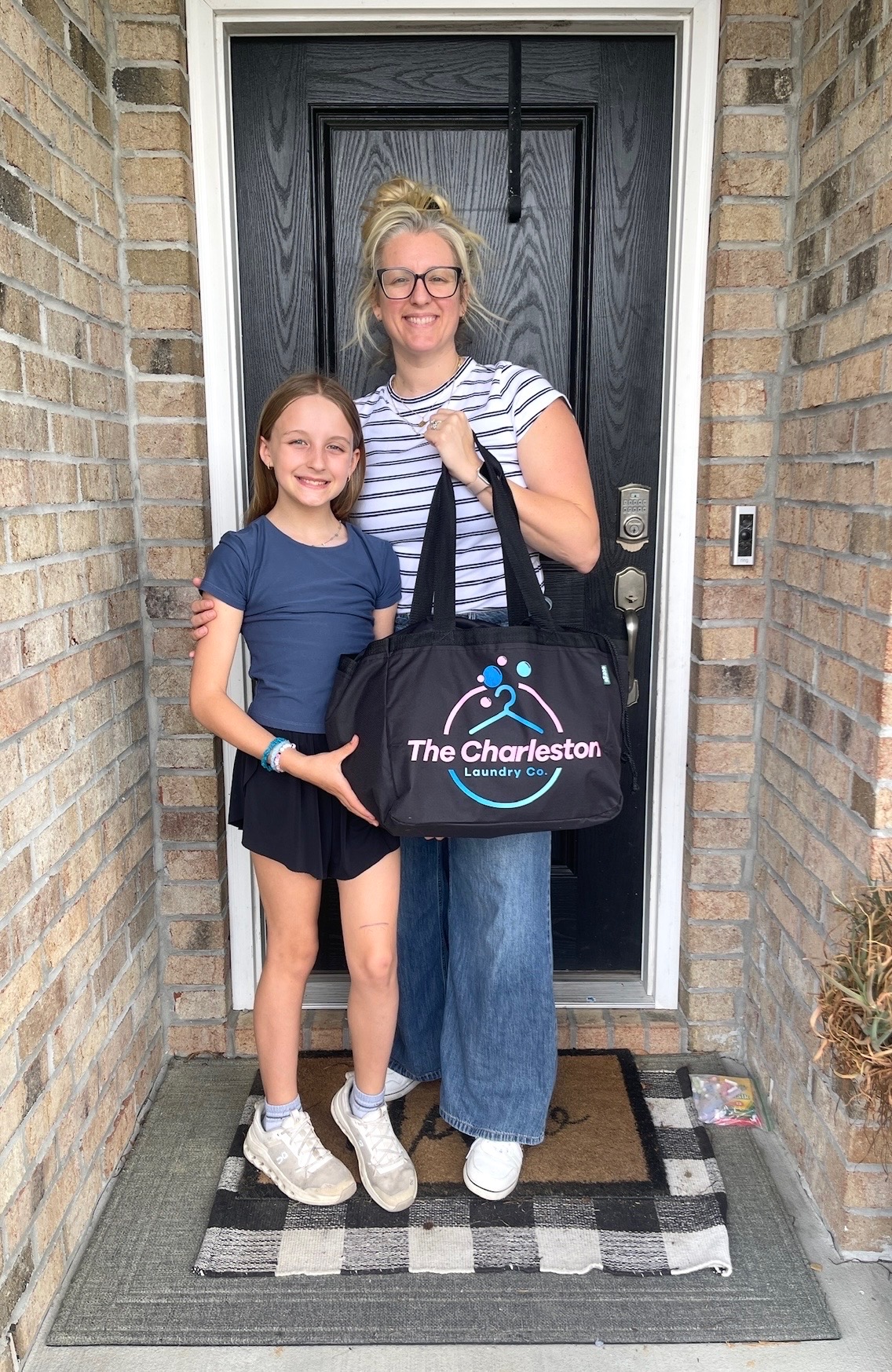 Two women stand in front of a black door with brick walls on sides, holding a black bag with blue and white design.