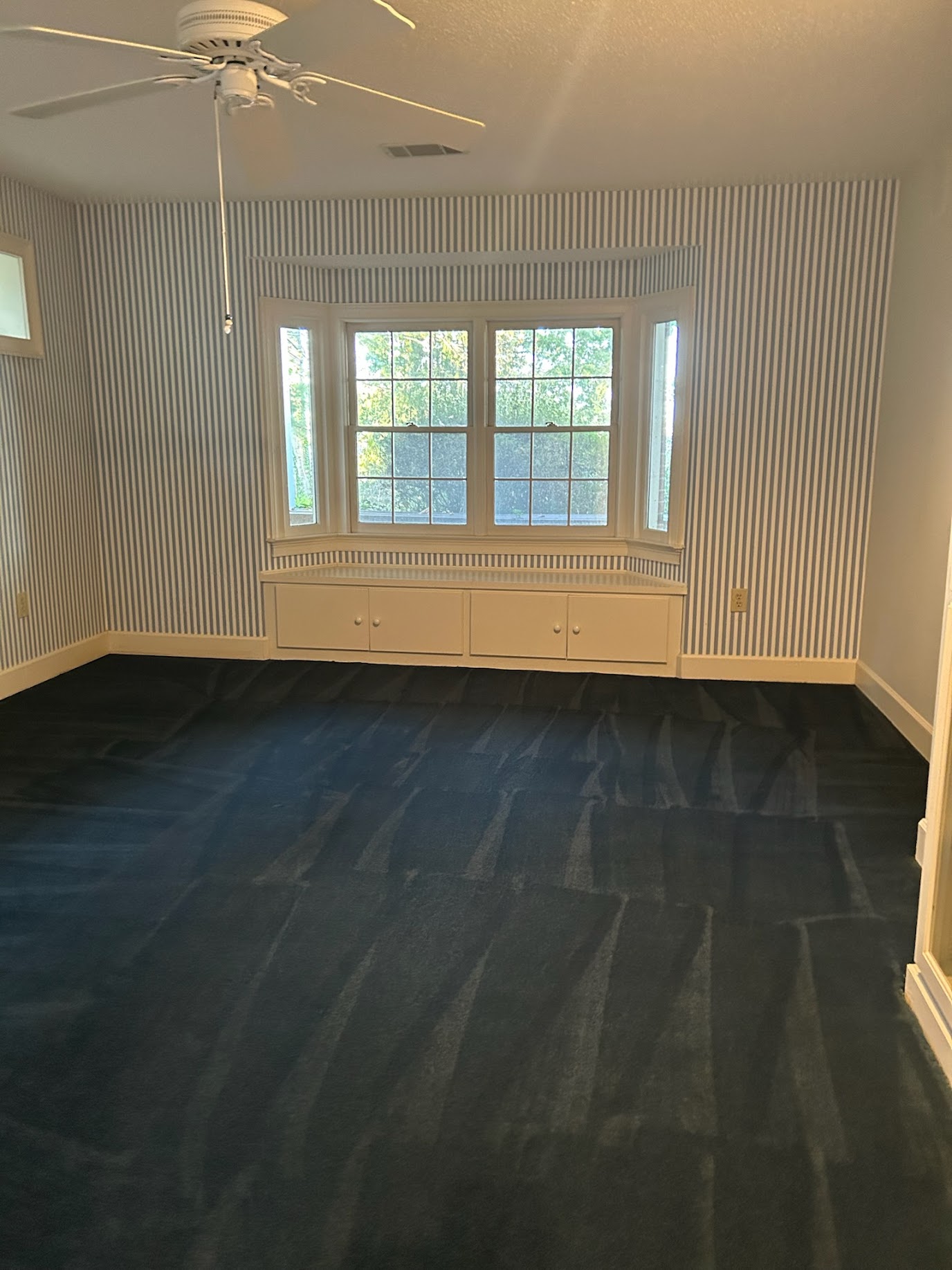 Empty room with large window, striped wallpaper, dark carpet, ceiling fan, and built-in window seat.