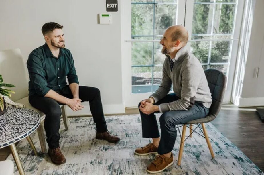 Two men sit and converse in a bright room with large windows and a patterned rug.