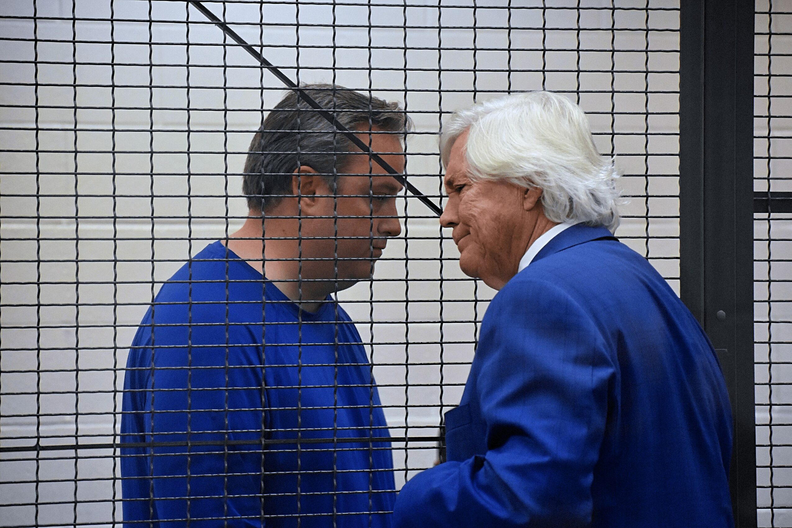 Two men face each other behind a wire mesh, one with dark hair and glasses, the other with white hair, both in blue suits.