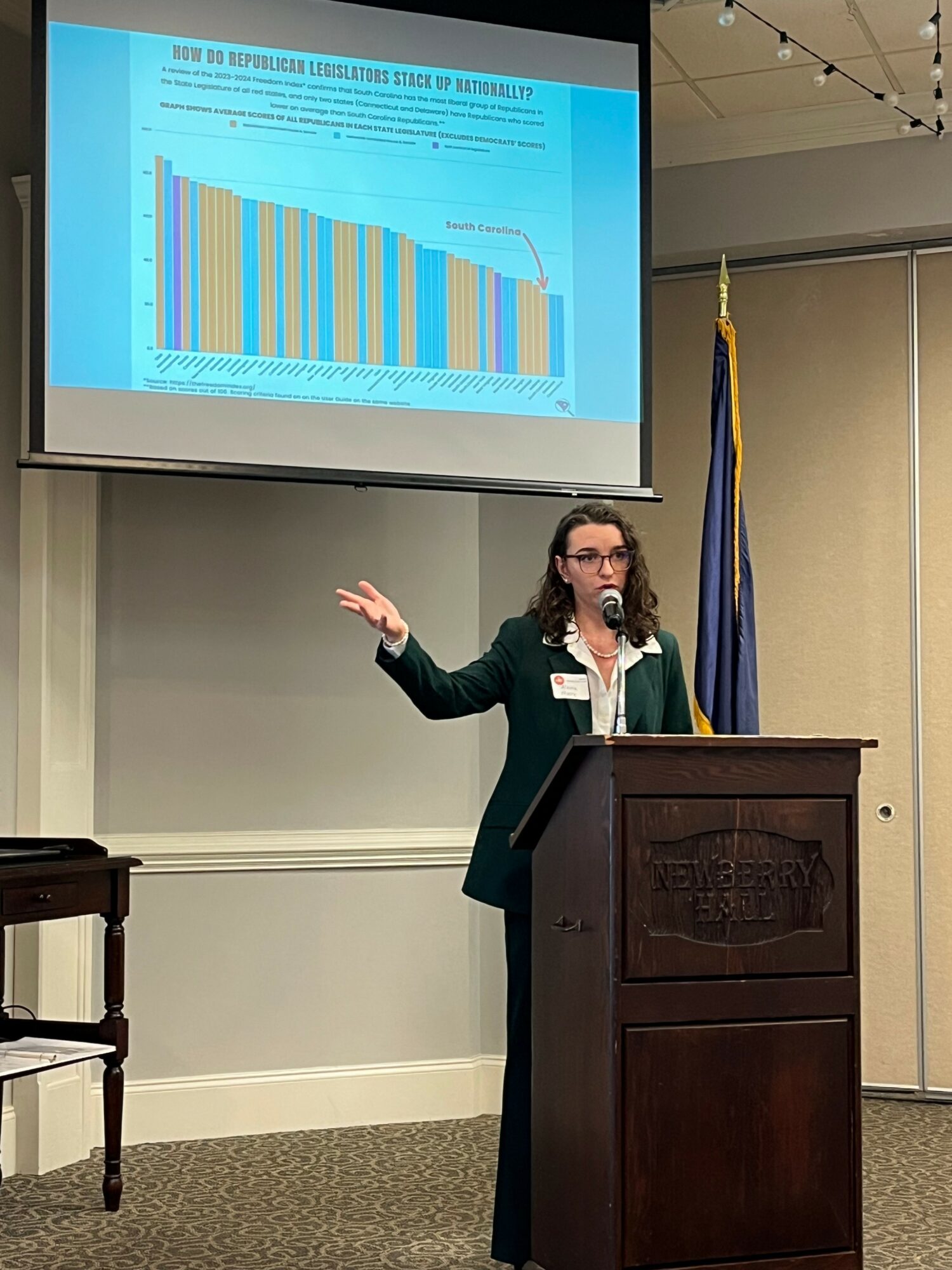 Woman in green blazer presenting at podium with a slide showing a bar graph projected behind her, in a conference room.