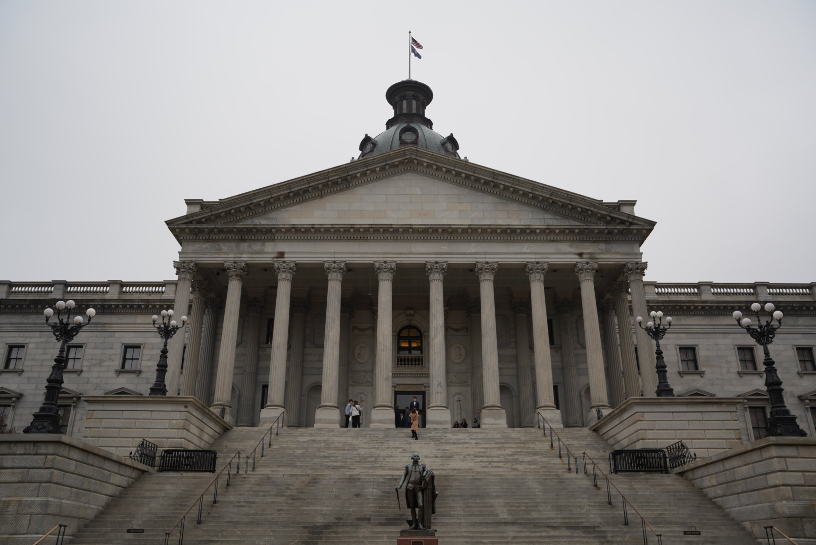 Front view of a classical government building with columns, stairs, and a flag on top, under an overcast sky.