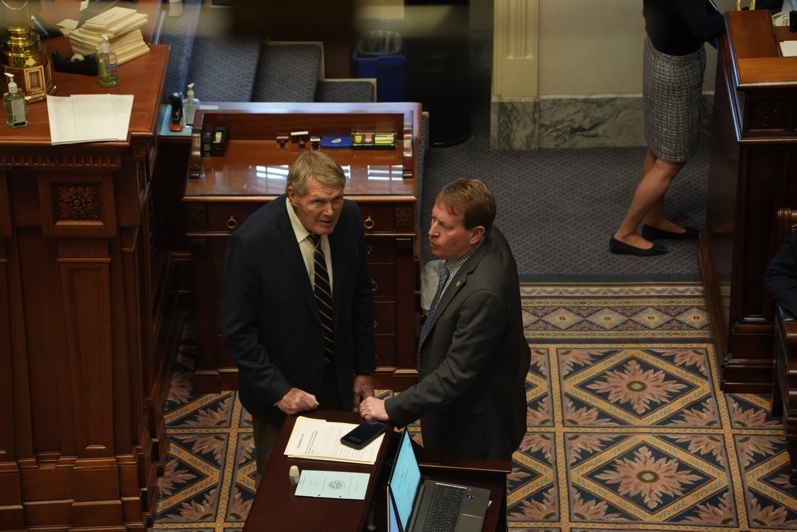 Two men in suits are talking in a formal room with wooden furniture and patterned carpet.