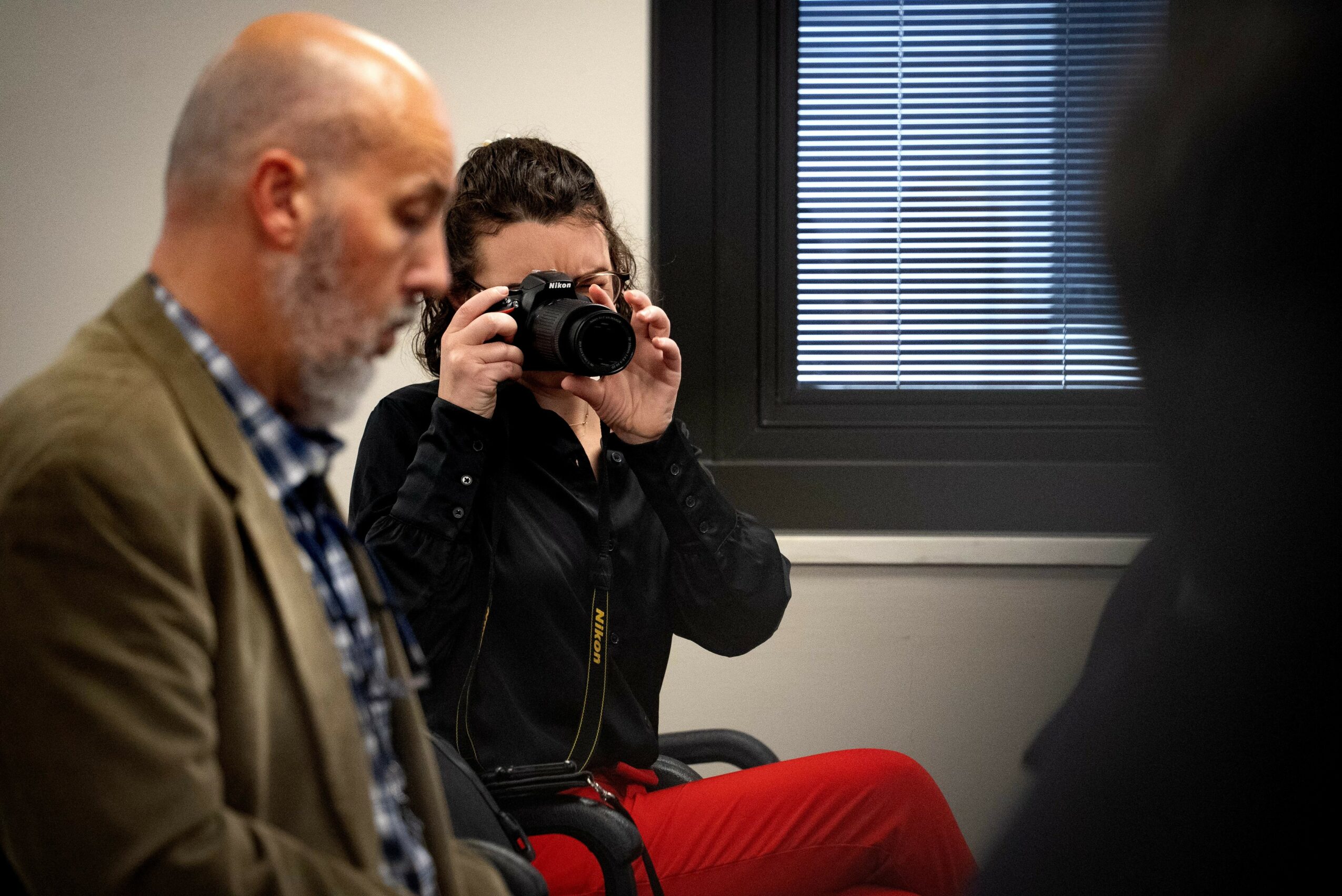 Person taking a photo with a camera in a room with a window and blinds, other people present, neutral setting.