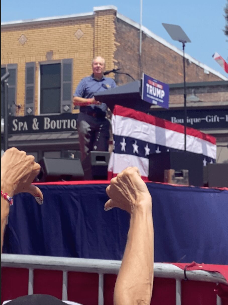 Man standing on a stage with a Trump sign, American flag-themed decorations, and a crowd in front, during a parade or rally.