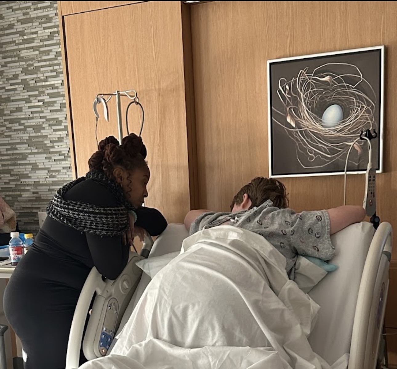 A person lying in a hospital bed with an IV, a woman leaning over, and a framed abstract artwork on the wall.