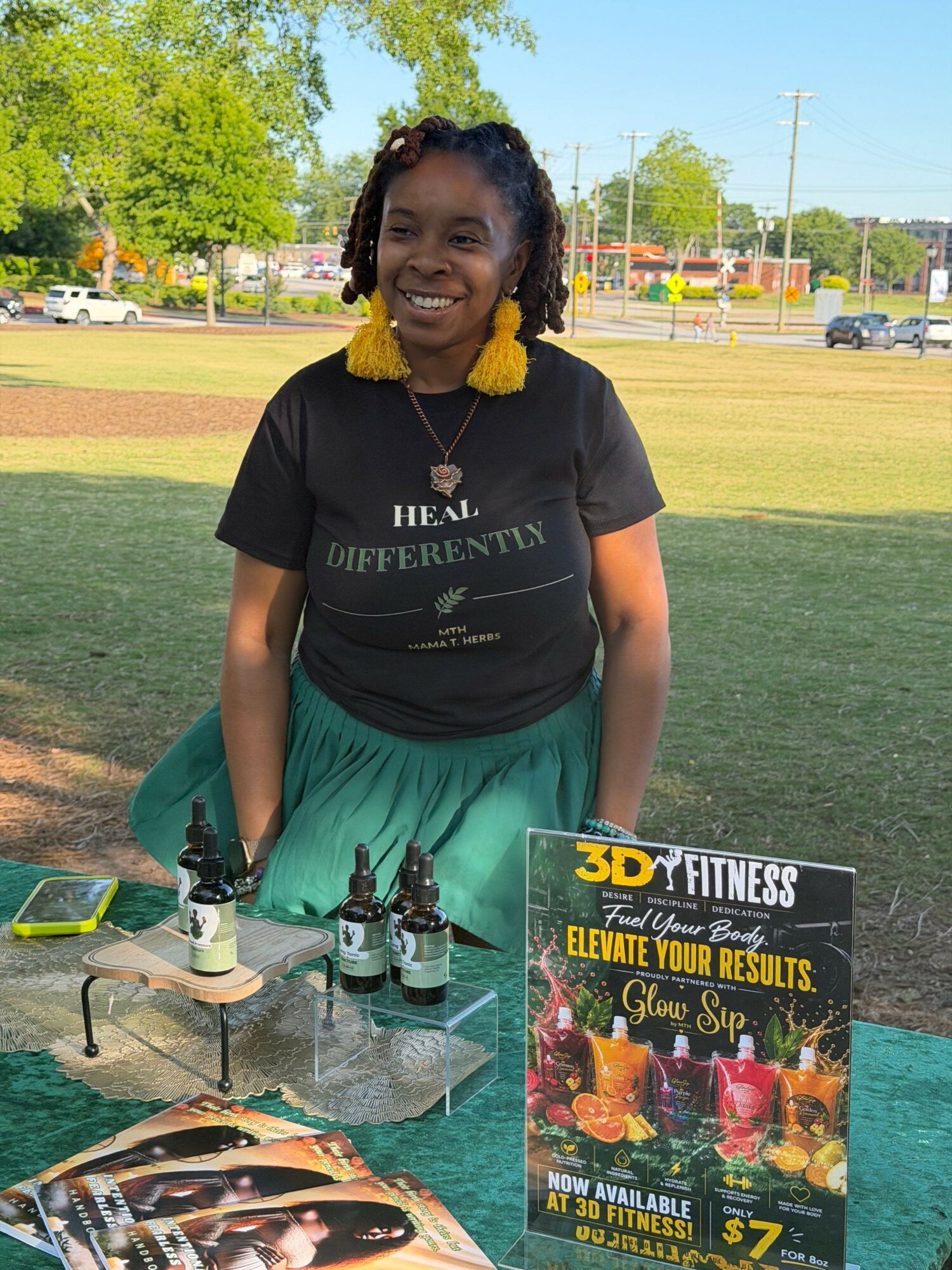 Young woman with braided hair and yellow earrings stands outdoors near a table with trophies and a poster, smiling.