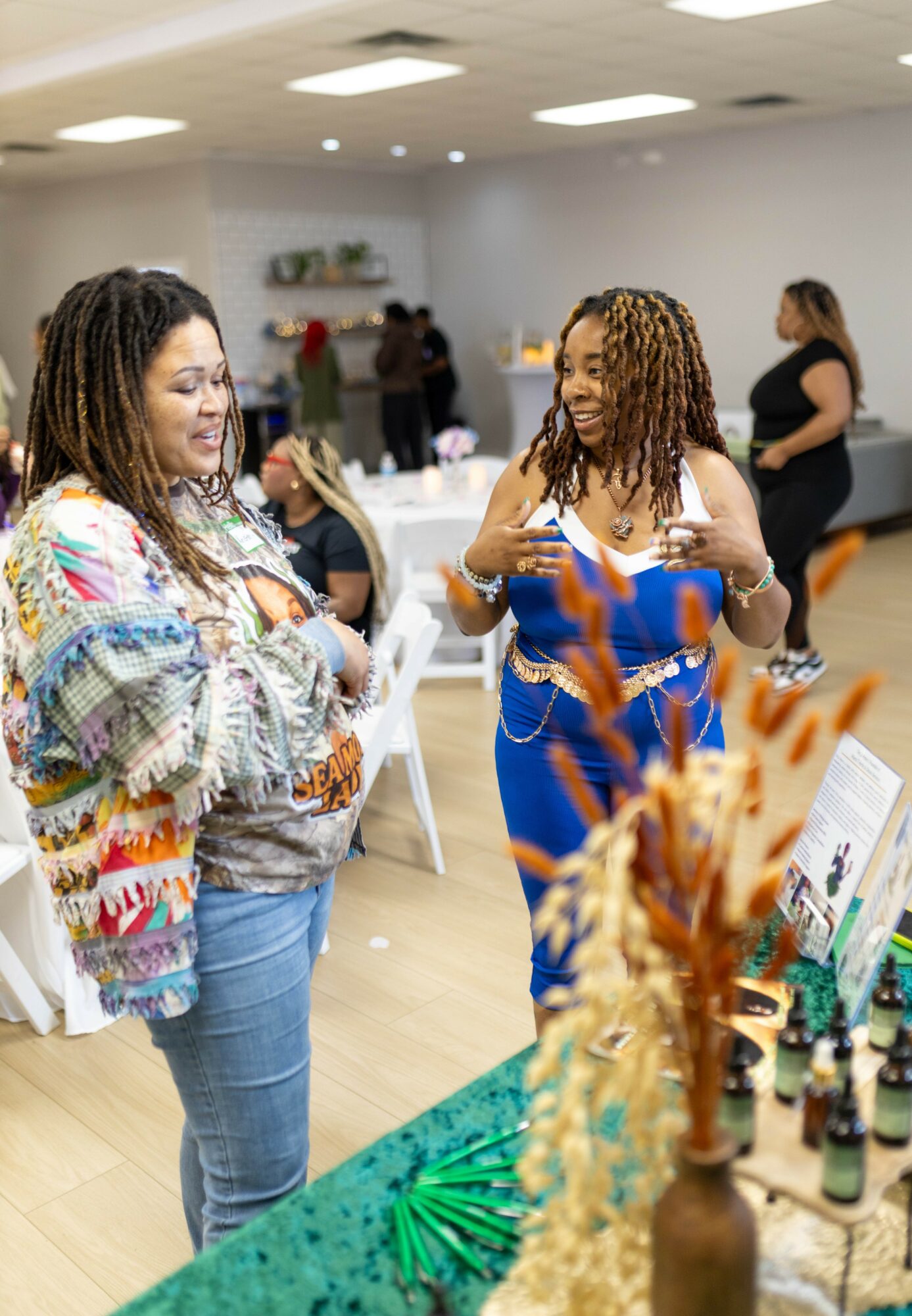 Two women with curly hair talking at an indoor event, with tables and other people in the background.