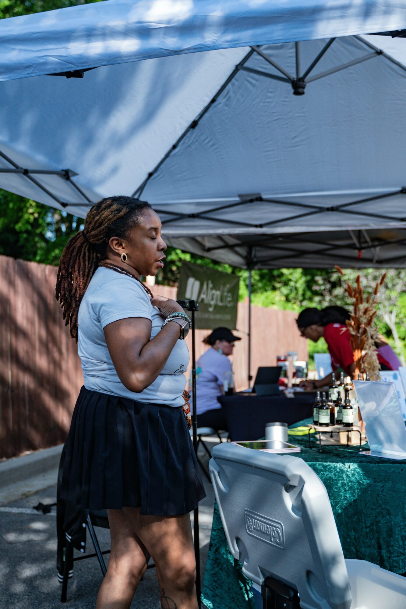 Woman with dreadlocks wearing a white shirt and black skirt standing under a canopy at an outdoor event.