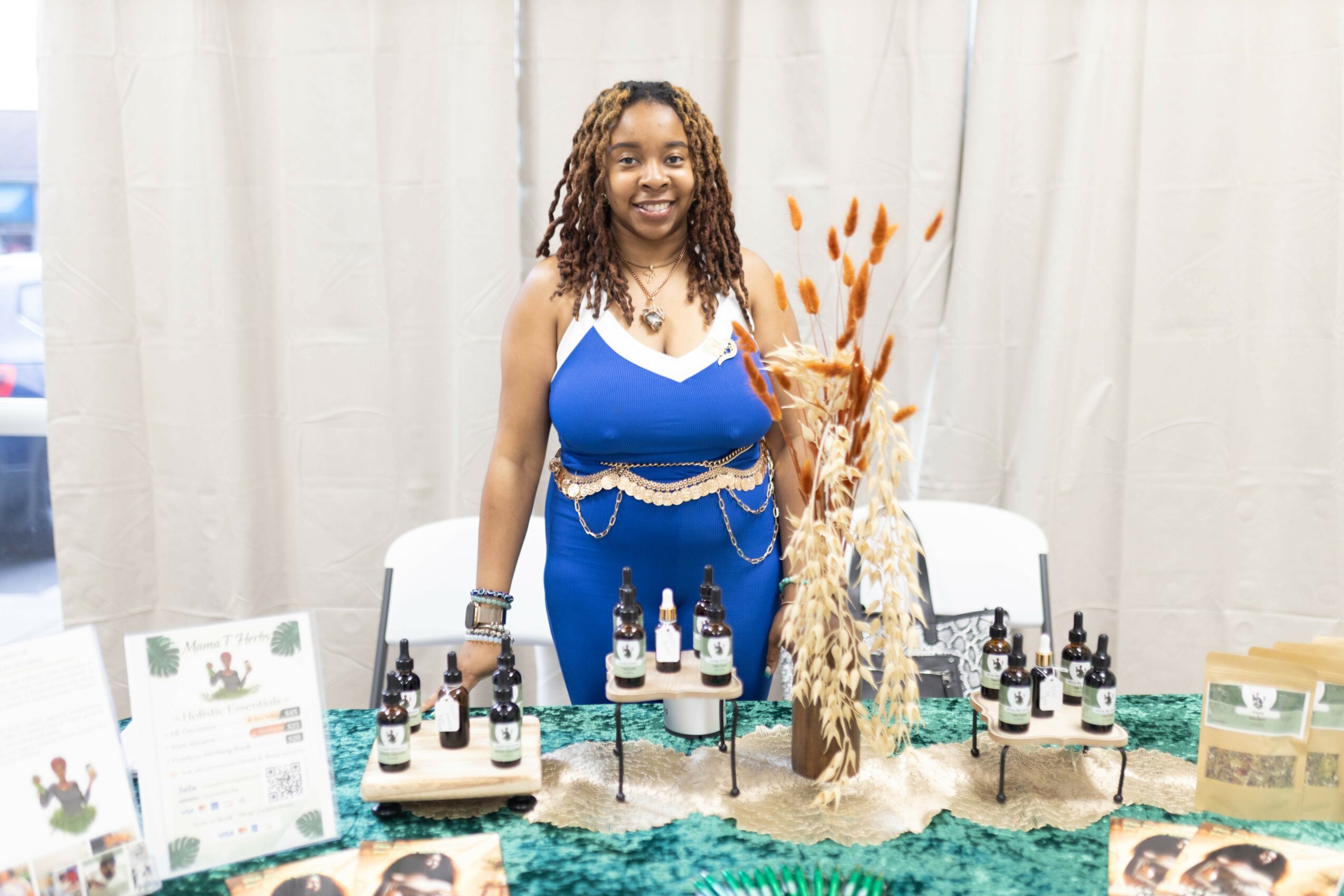Smiling woman in blue dress standing behind a table with essential oils and products, beige curtain background.