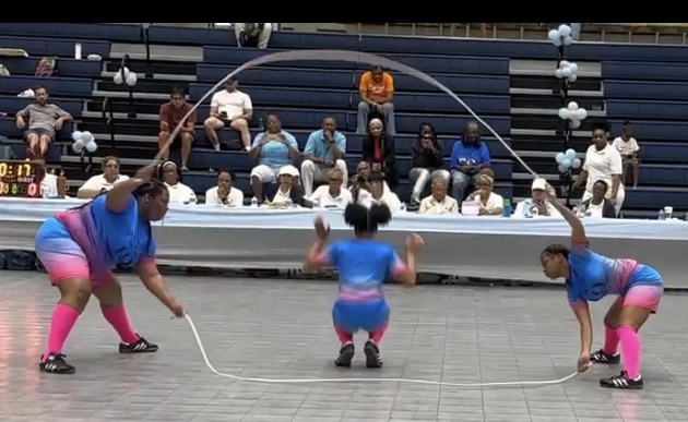 Three performers in colorful outfits jump rope in an indoor arena with an audience seated in the background.