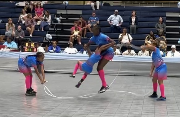 Four children in colorful outfits jump rope in a gymnasium with spectators watching, some seated on bleachers.