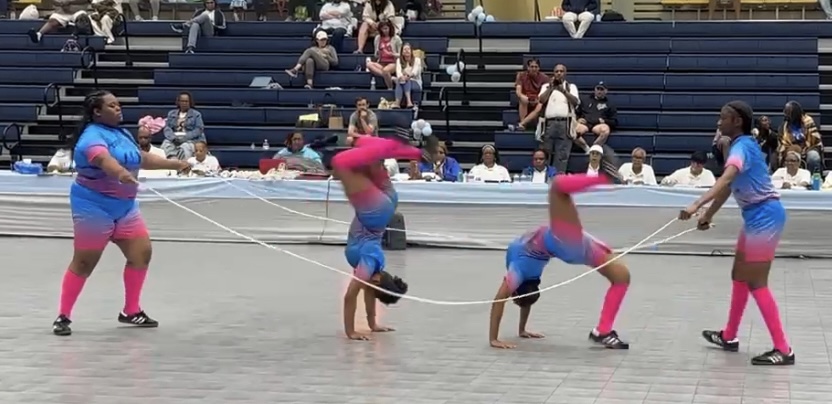 Four performers in colorful costumes are engaged in a synchronized ribbon routine on a gymnasium floor with an audience in the background.