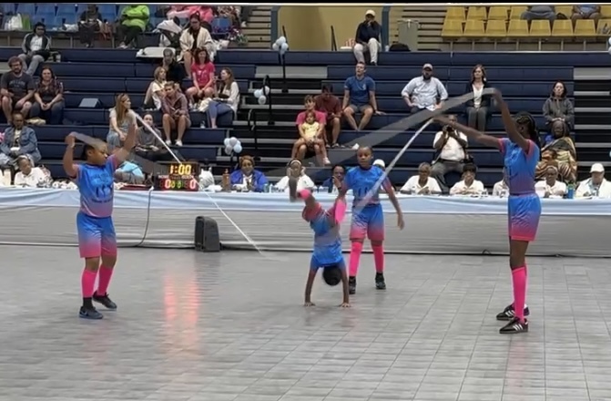 Four performers in colorful costumes jump rope in an indoor arena with seated audience and spectators in the background.