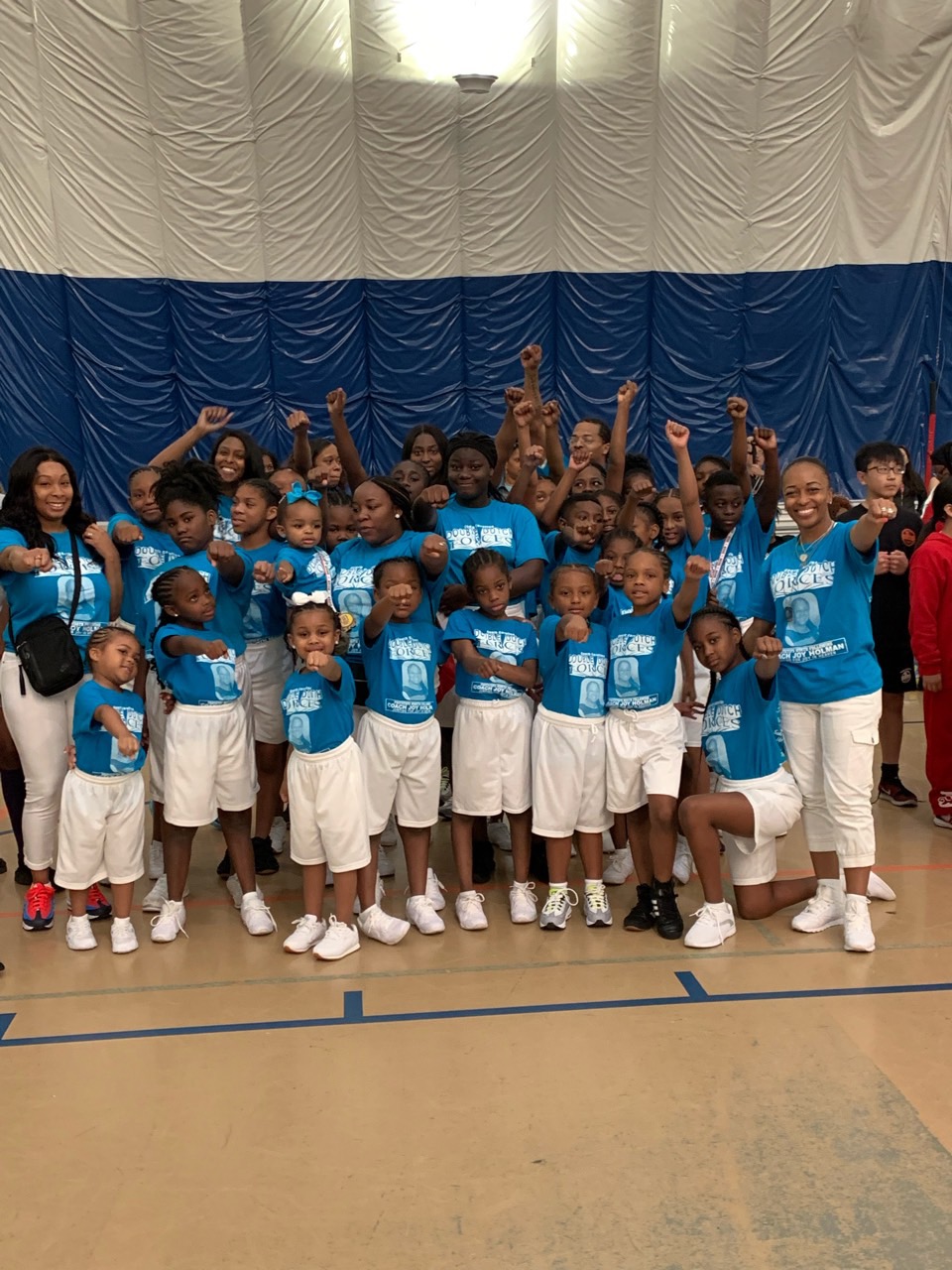 Group of children and adults in matching blue shirts and white shorts posing indoors, some raising fists, in front of a blue and white curtain.