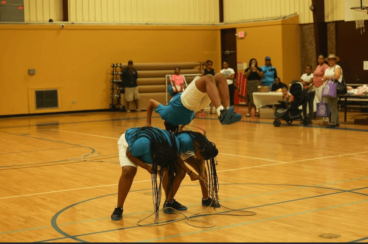 People playing basketball in a gymnasium with a yellow wall and wooden floor, some sitting and others standing in the background.