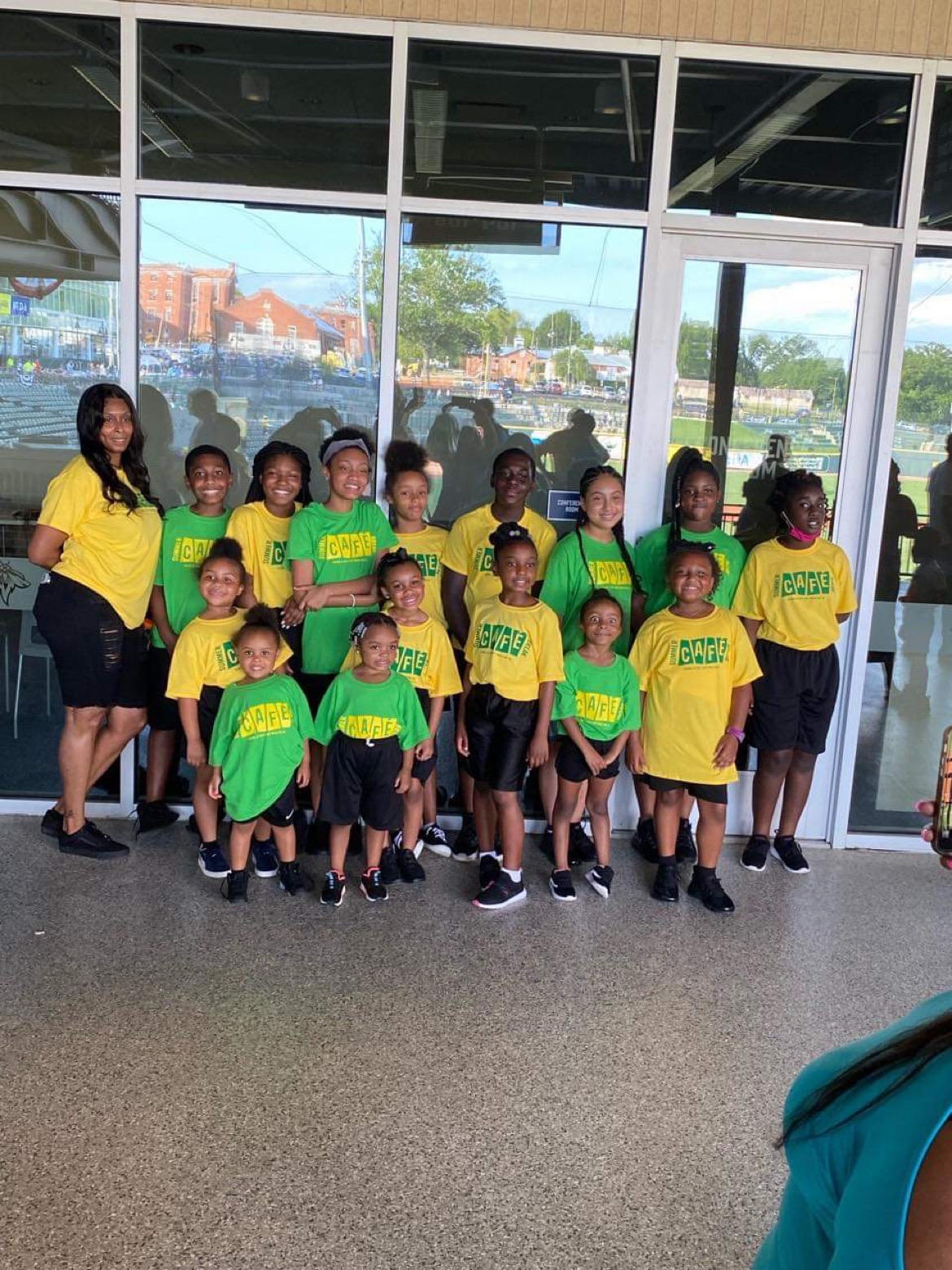 Group of children and adults in yellow and green shirts standing inside near glass doors, with a cityscape outside.