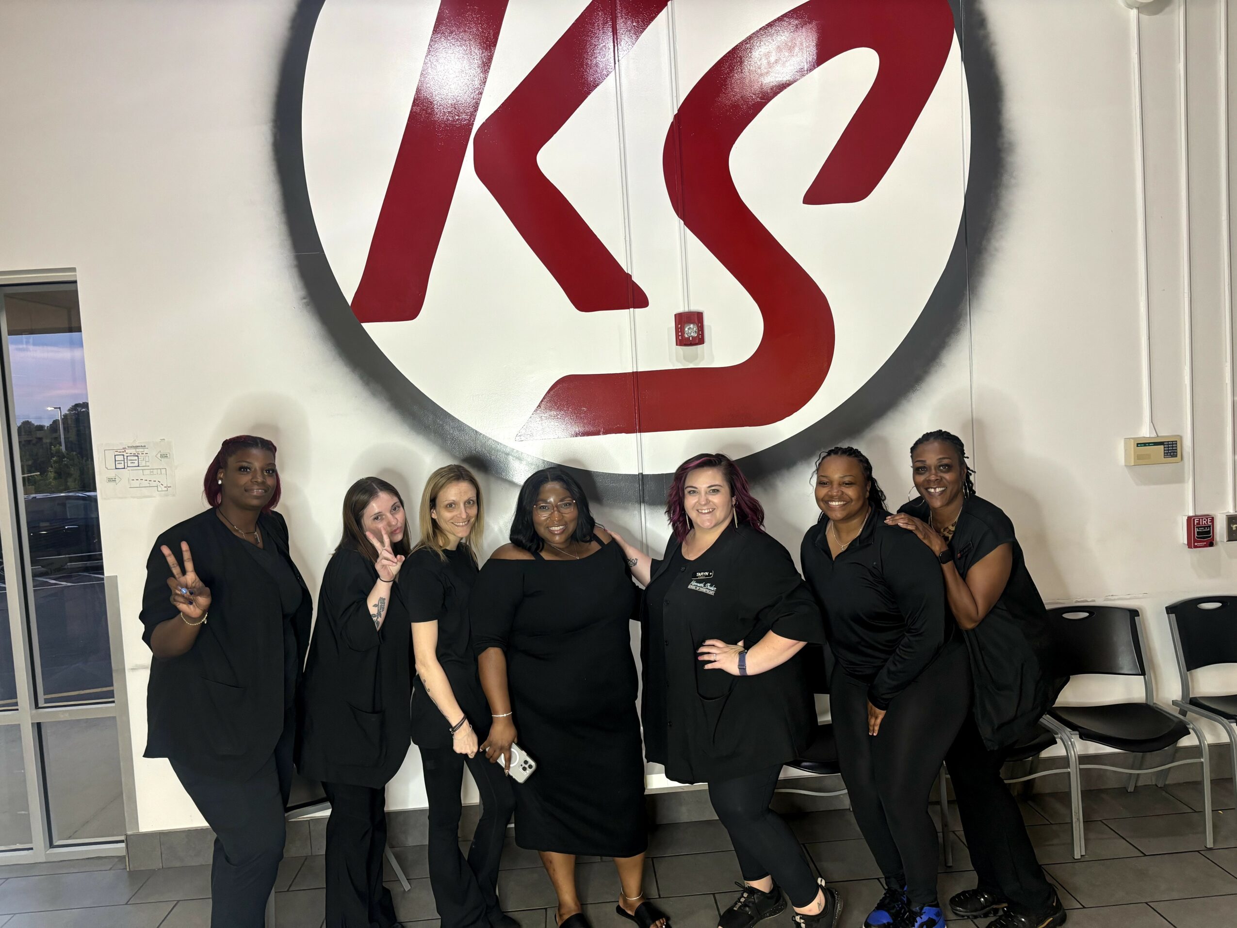 Six women stand in front of a large circular logo with red and black design on a white wall, smiling and posing.