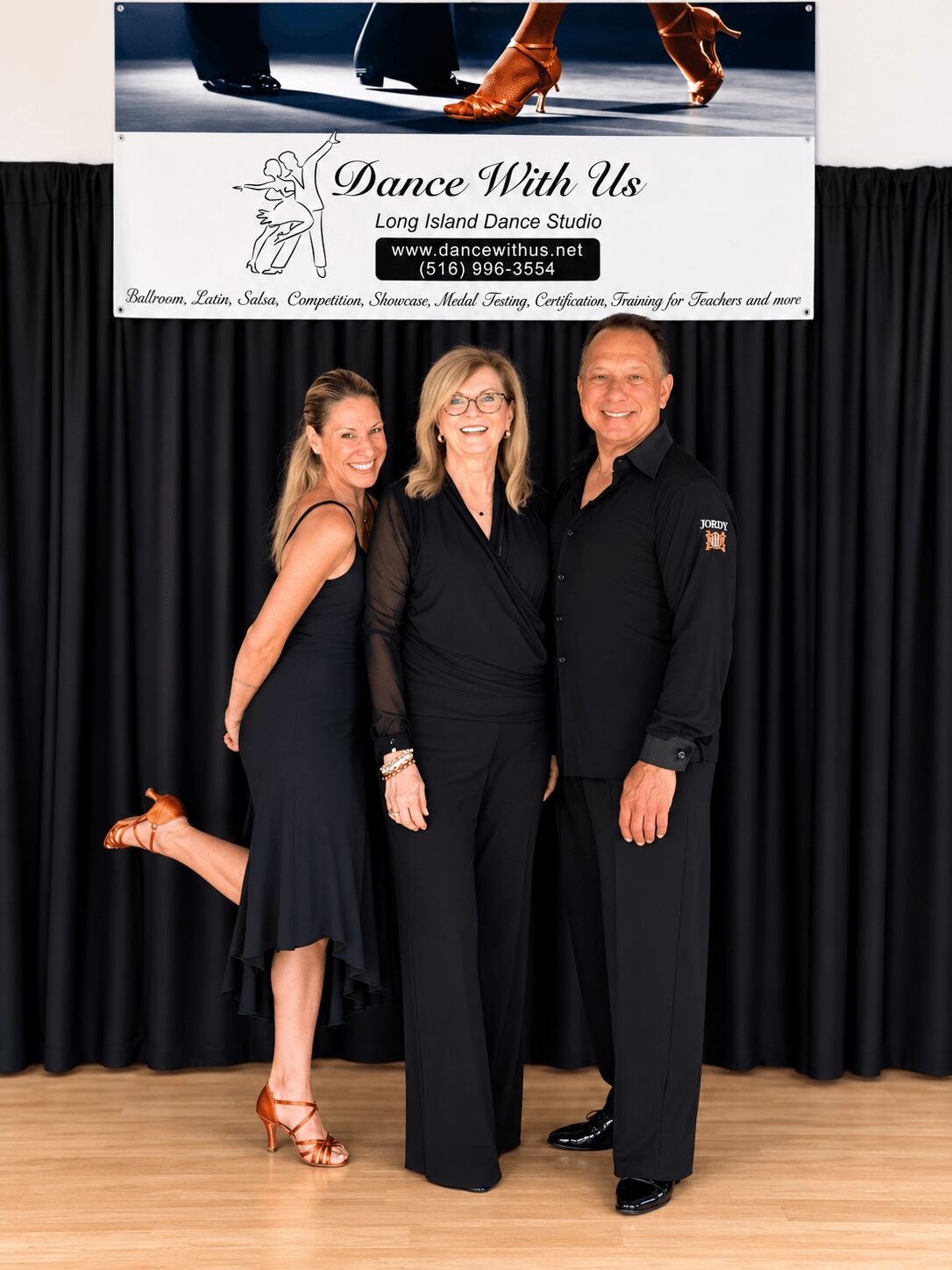 Three people standing on stage in front of a black curtain, smiling, with a dance studio banner above them.