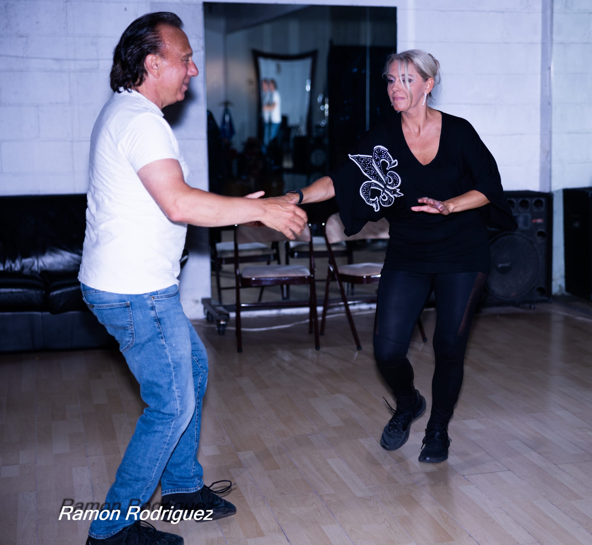 Two people dancing and smiling, one holding a trophy, in a room with wooden floor and chairs, woman clapping.