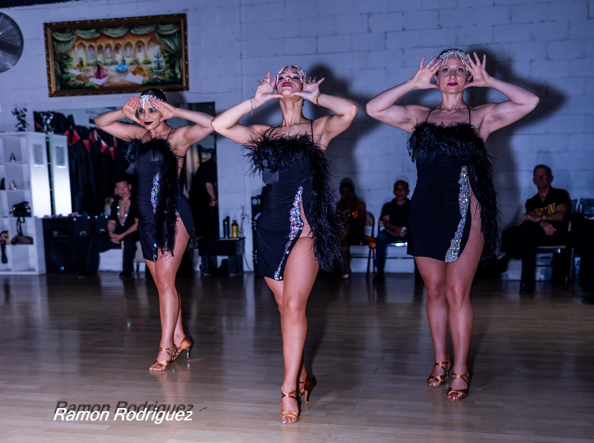 Three dancers in black costumes perform a synchronized dance with hands near their faces, in a room with a painting and seated audience.