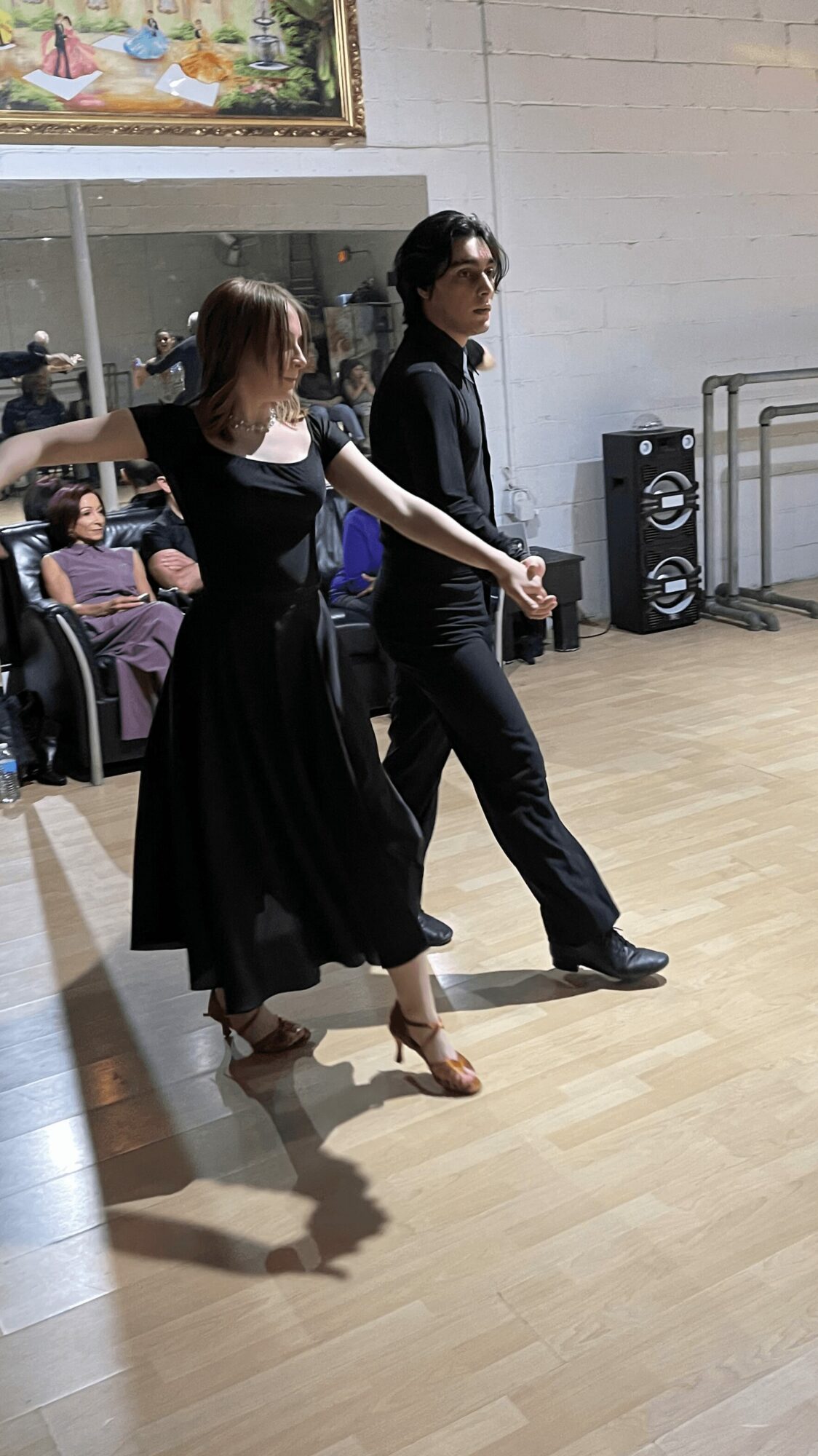 A couple dancing in a room with a wooden floor, with an audience watching through a window in the background.