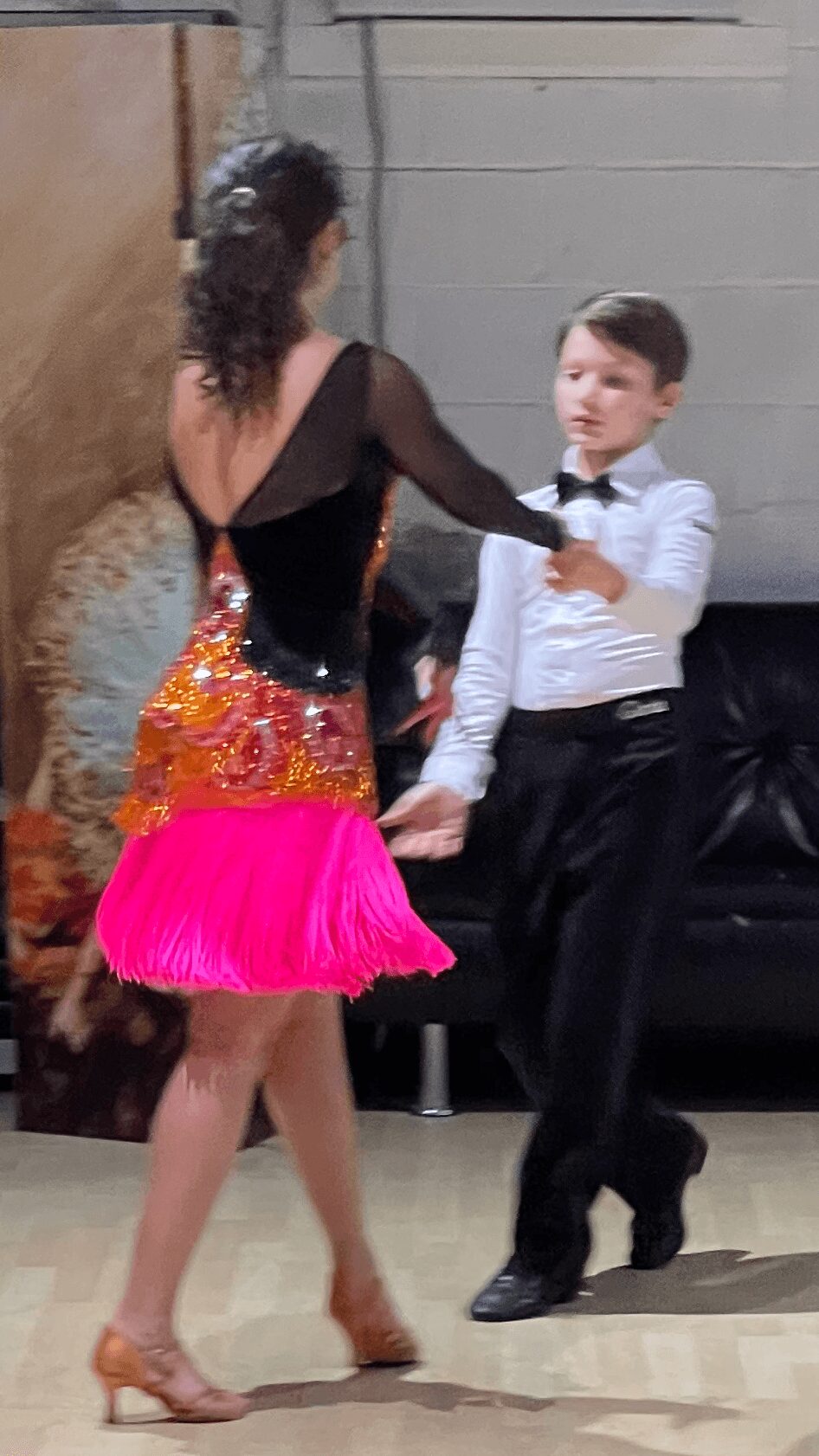 A woman and a boy dance indoors, woman in colorful dress, boy in white shirt and black pants, holding hands.