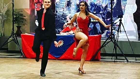 Two performers, one in a suit and one in a red costume, dance on stage with a decorated table behind them.