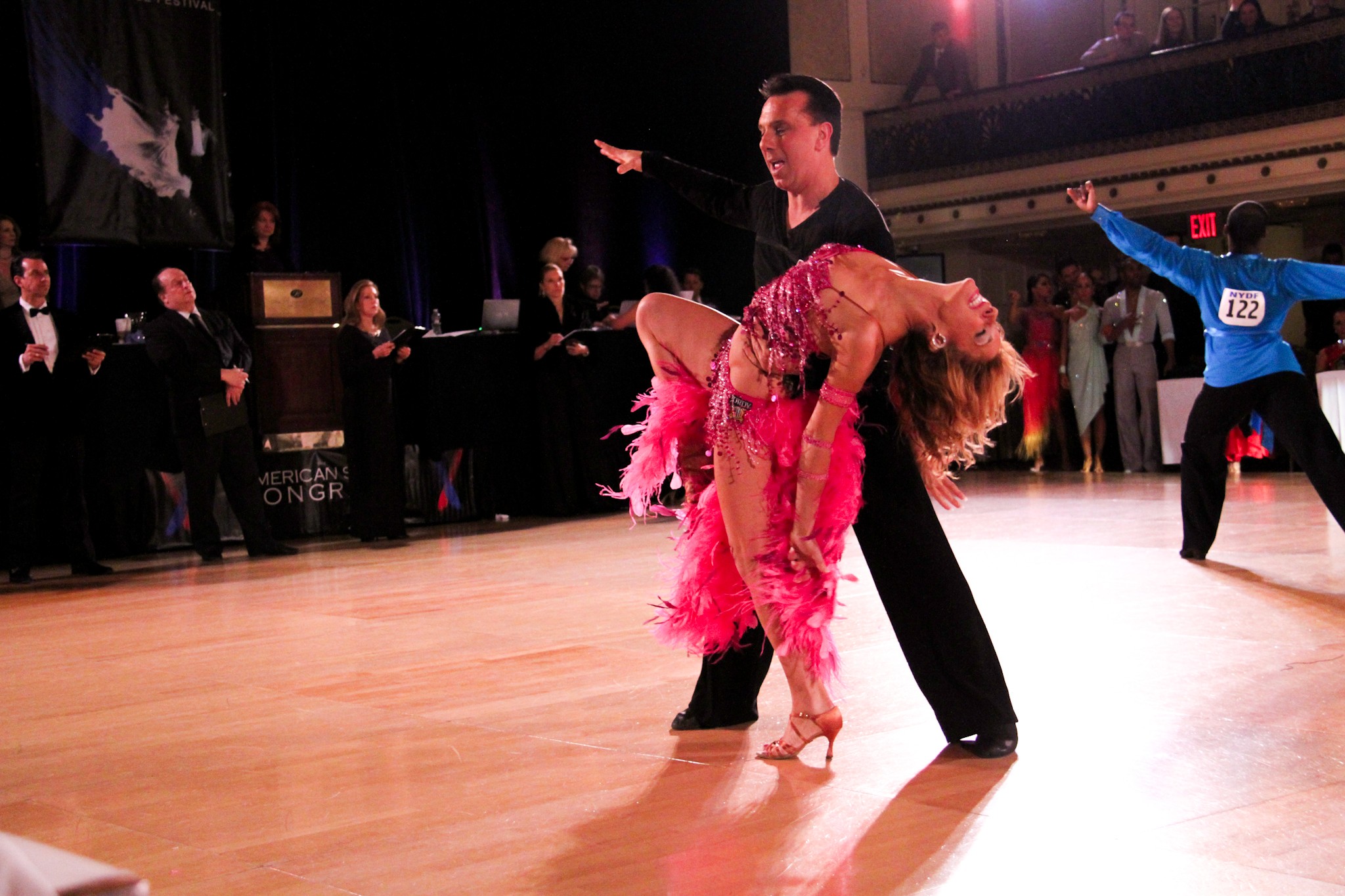 A couple dancing on a wooden floor, with the woman in a pink dress and the man in black, in a ballroom setting.