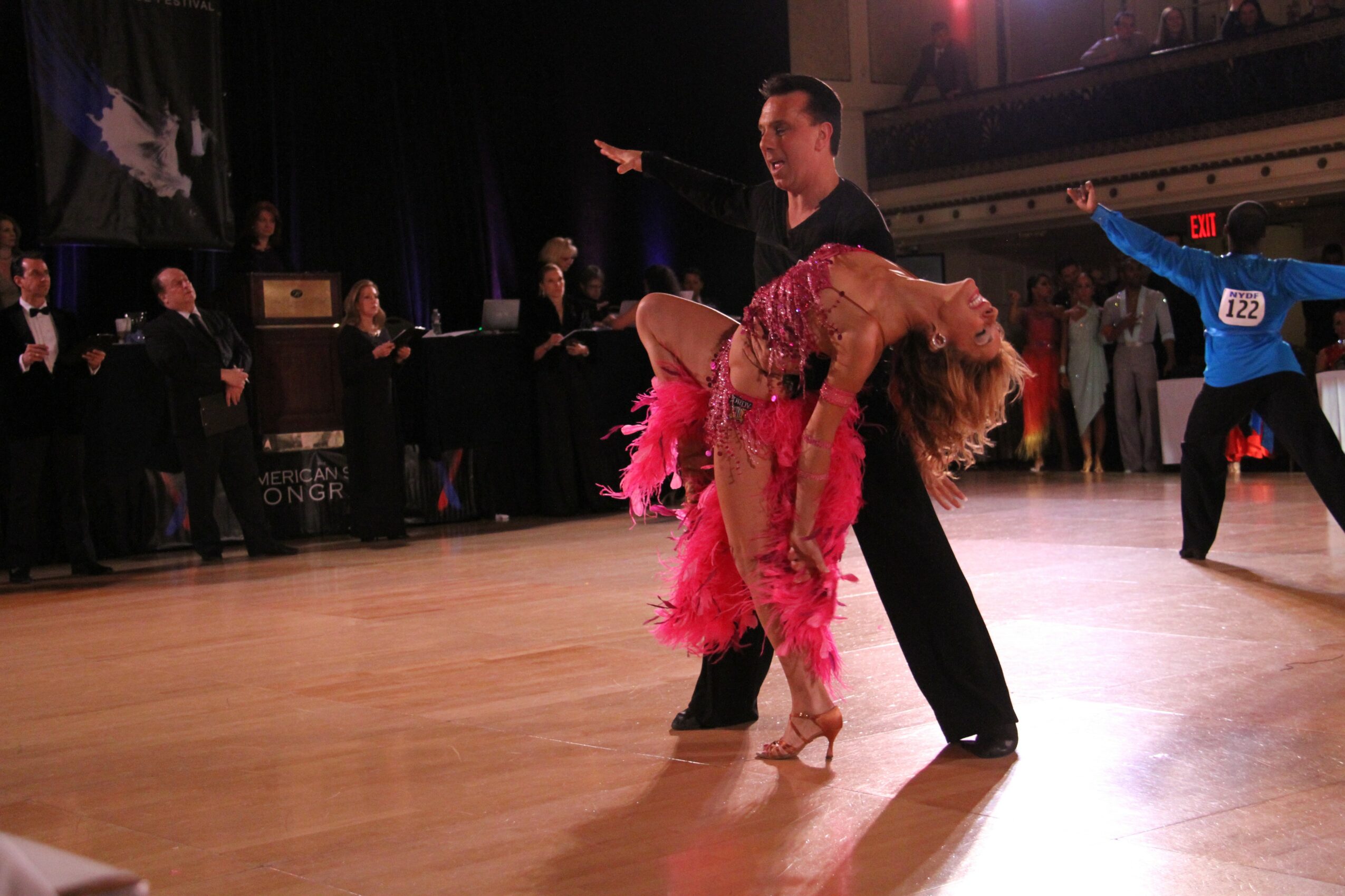 A couple dancing on a wooden floor with spectators and judges watching in the background.