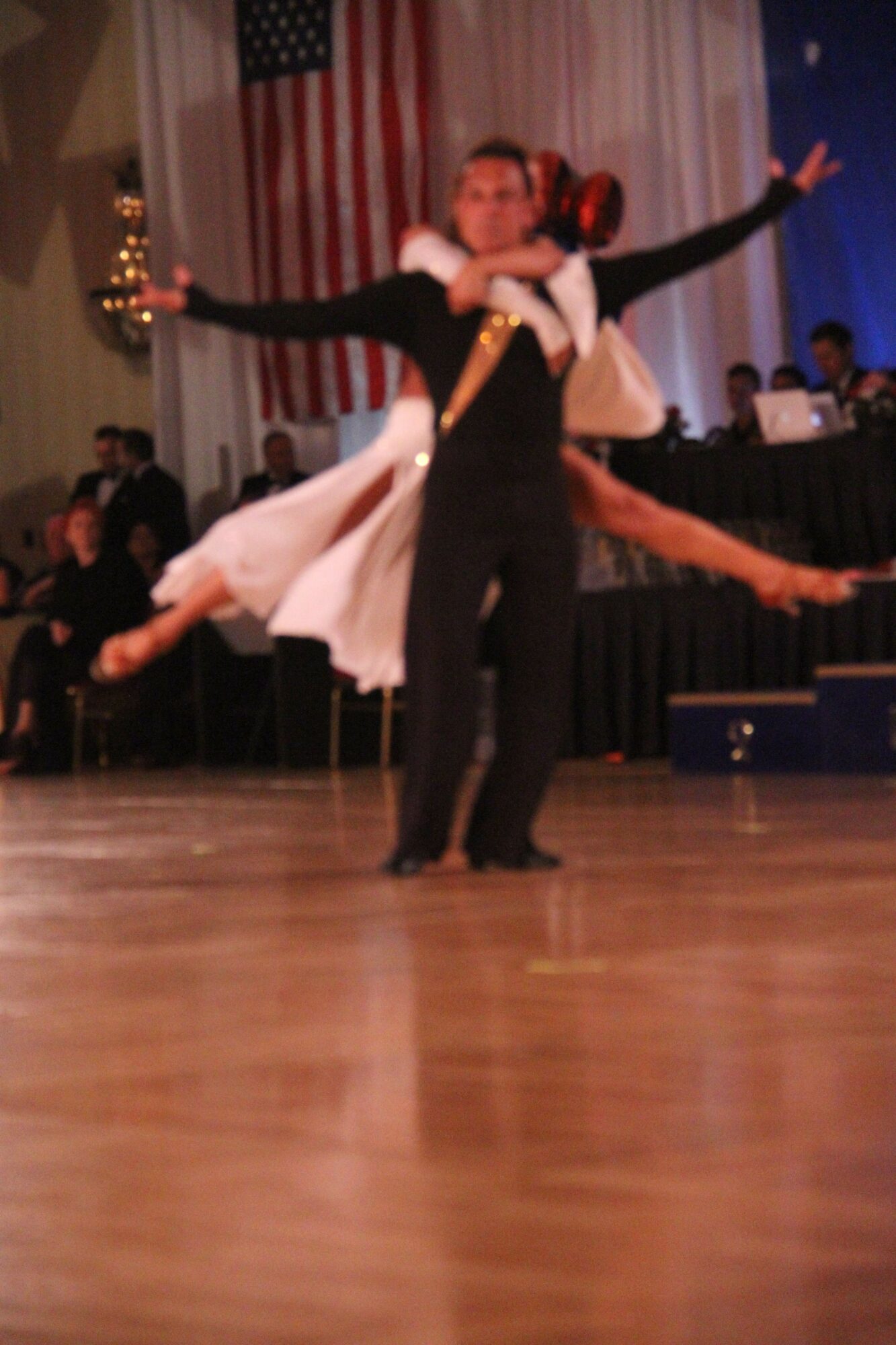 Person dancing on stage with arms outstretched, wearing a white dress and black jacket, American flag in background.