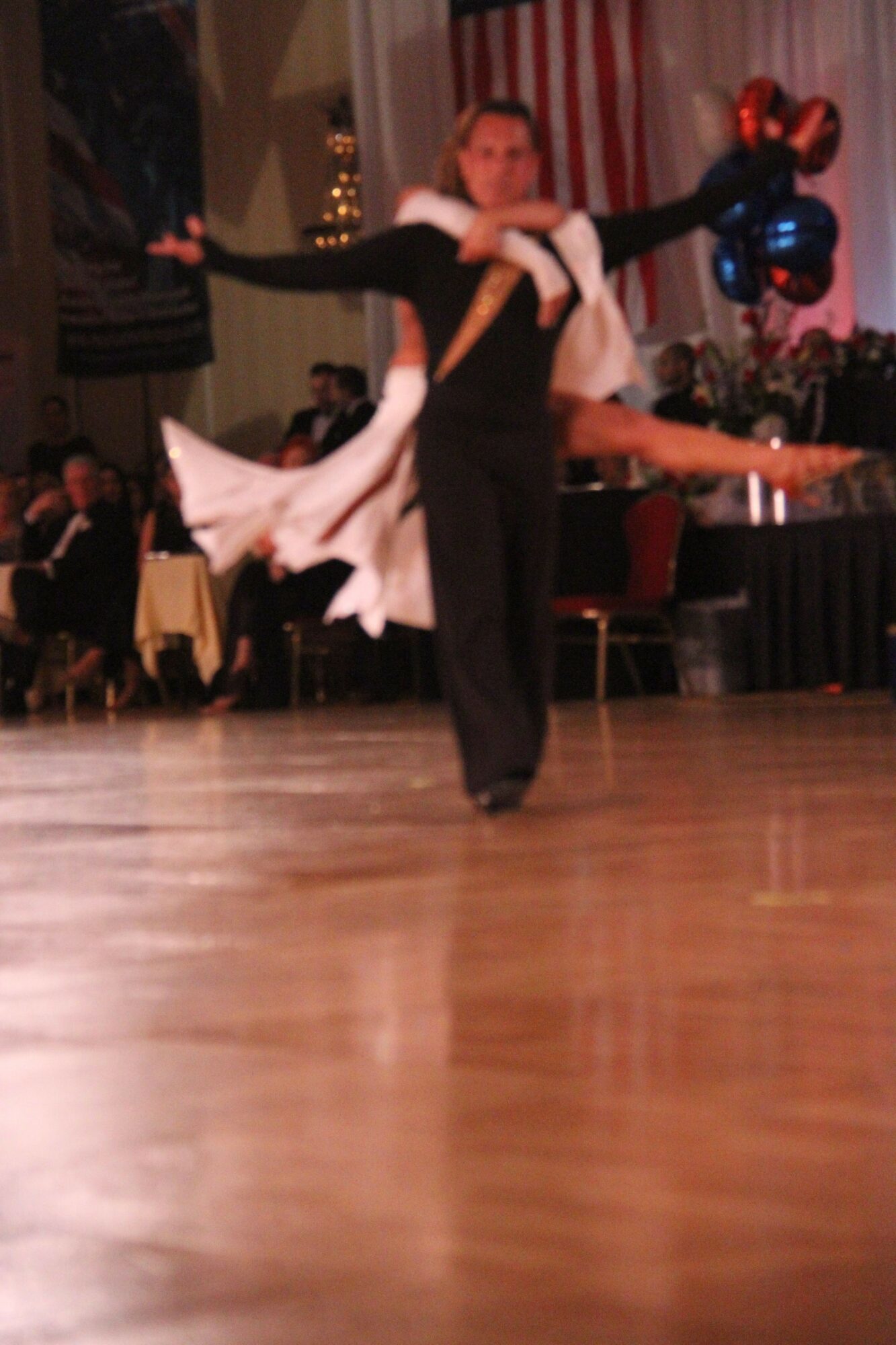 Person performing a dance move on stage with an audience and balloons in the background.