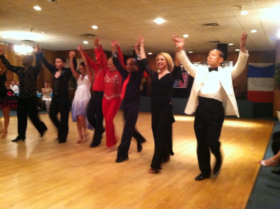 Group of people dancing in a line with hands raised, in a room with wooden floor and ceiling lights.