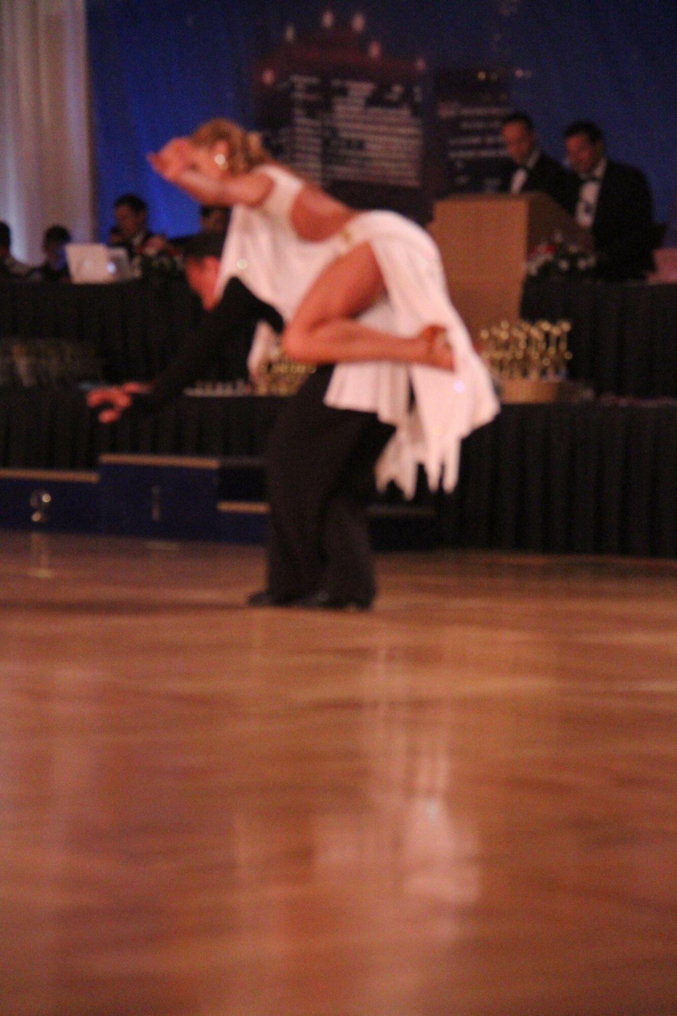 A couple dancing on a wooden floor, with a pianist and other people in the background, in a dimly lit room.