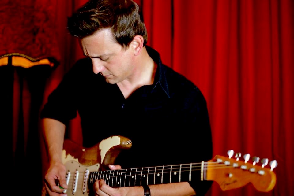Person playing electric guitar with red curtain background, looking down at the instrument.