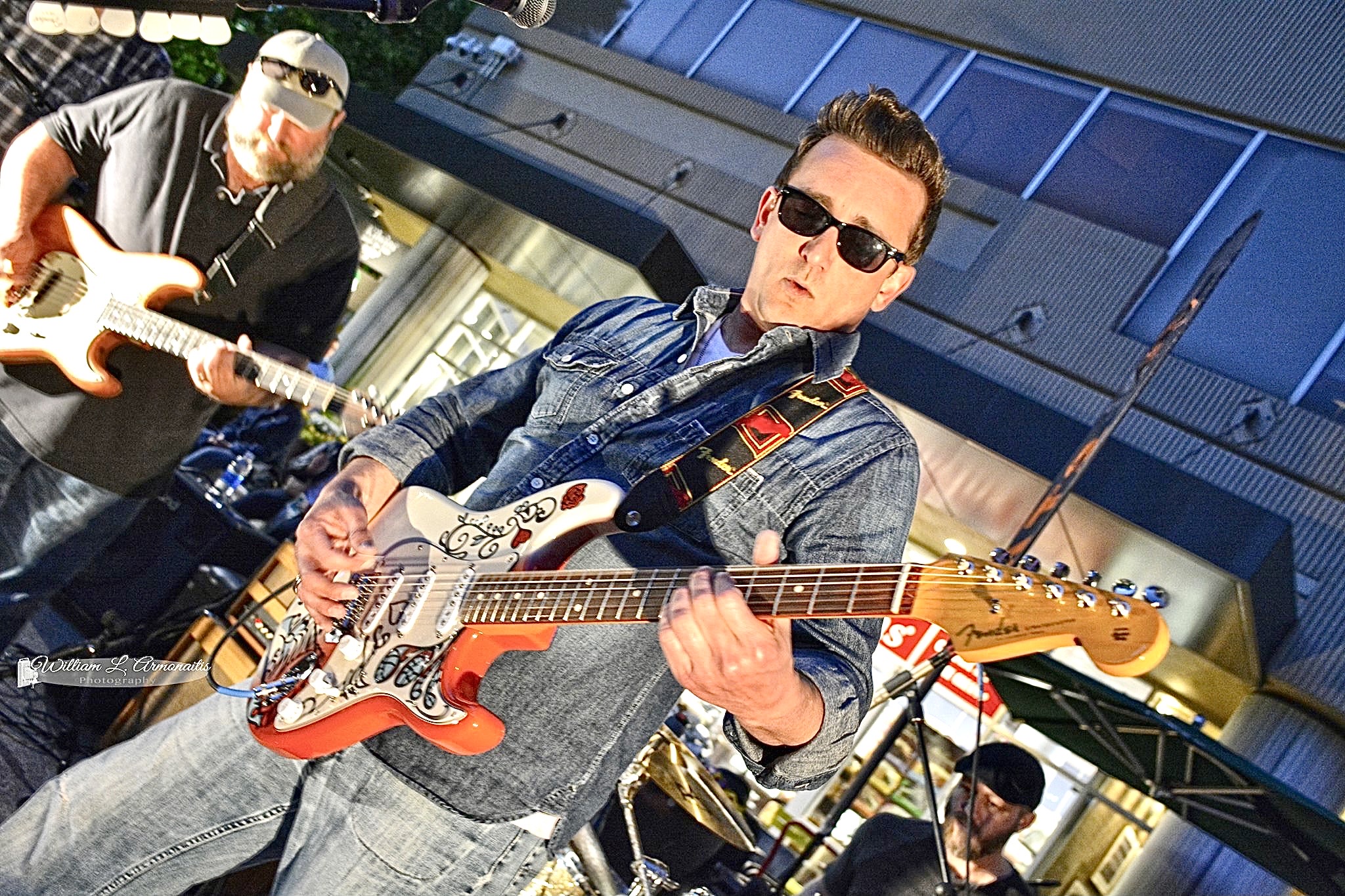 Two musicians playing electric guitars outdoors, one wearing sunglasses and denim jacket, the other in black, with a building background.