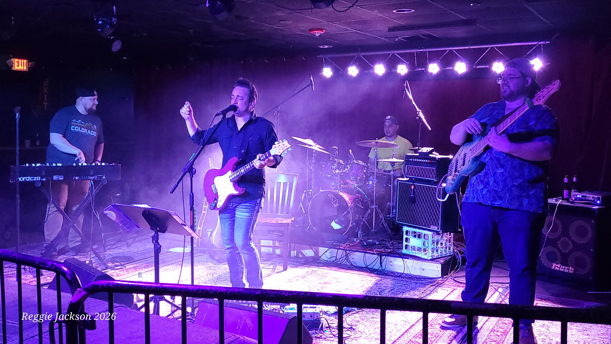 Band performs on stage with purple lighting, four musicians playing instruments, and a keyboardist on the left.