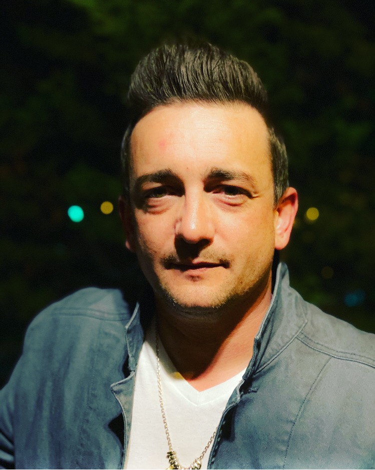 Man with short dark hair and light skin, wearing a gray jacket and white shirt, outdoors at night.