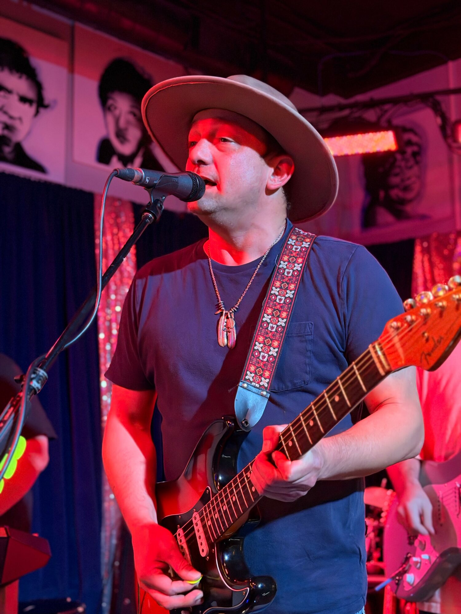 Male musician singing into microphone while playing electric guitar, wearing a wide-brimmed hat and dark shirt, in a colorful venue.