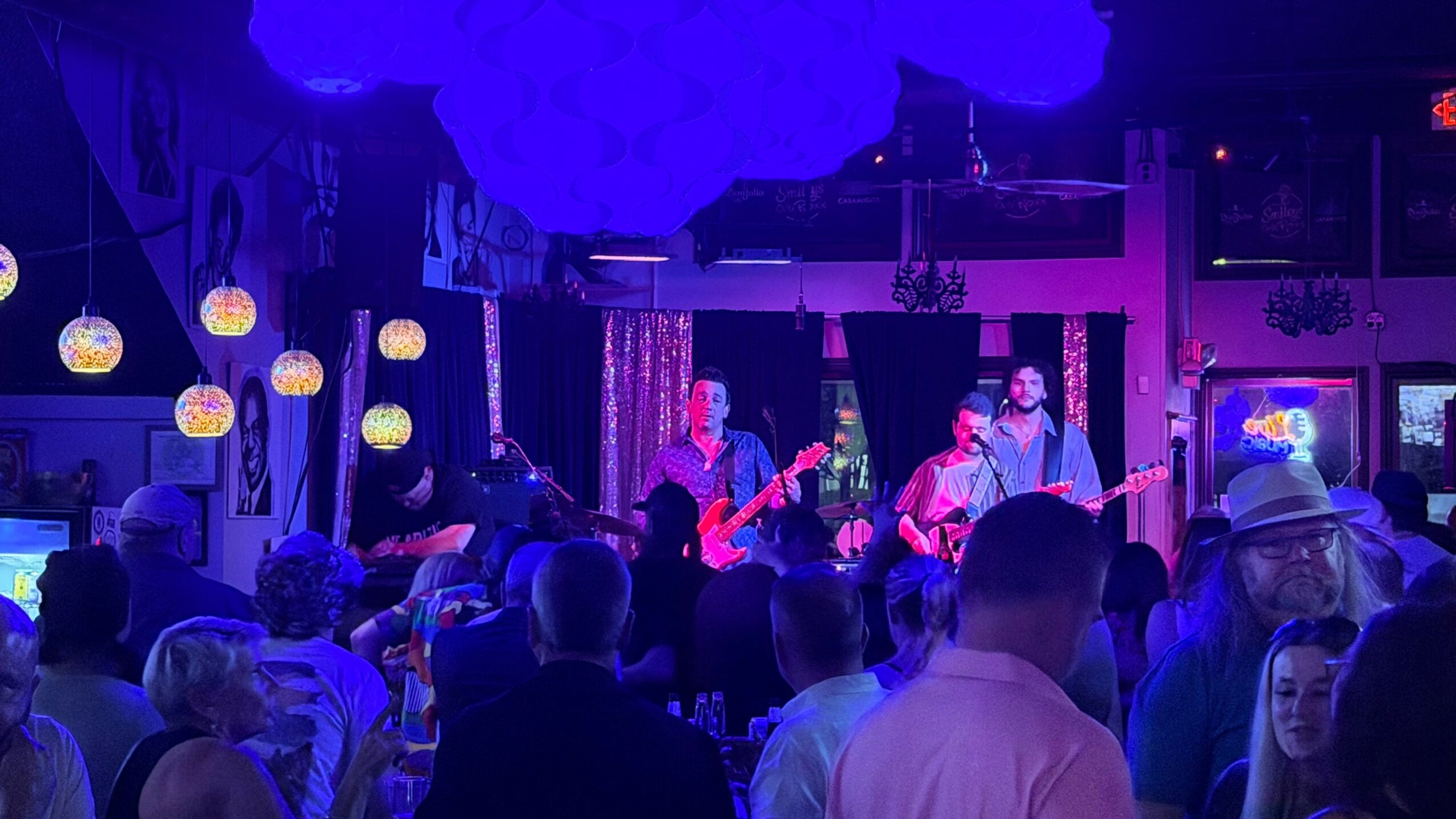 Band performing on stage with audience watching in a dimly lit venue, blue lighting, and hanging decorations.