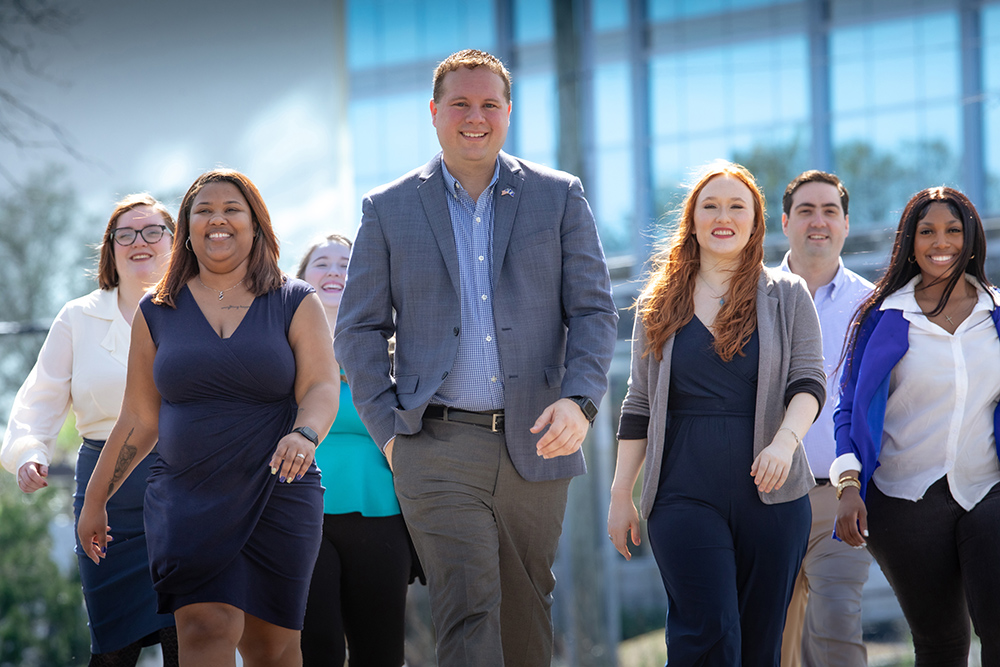 Group of diverse professionals walking together indoors, smiling, with large windows and modern architecture in background.