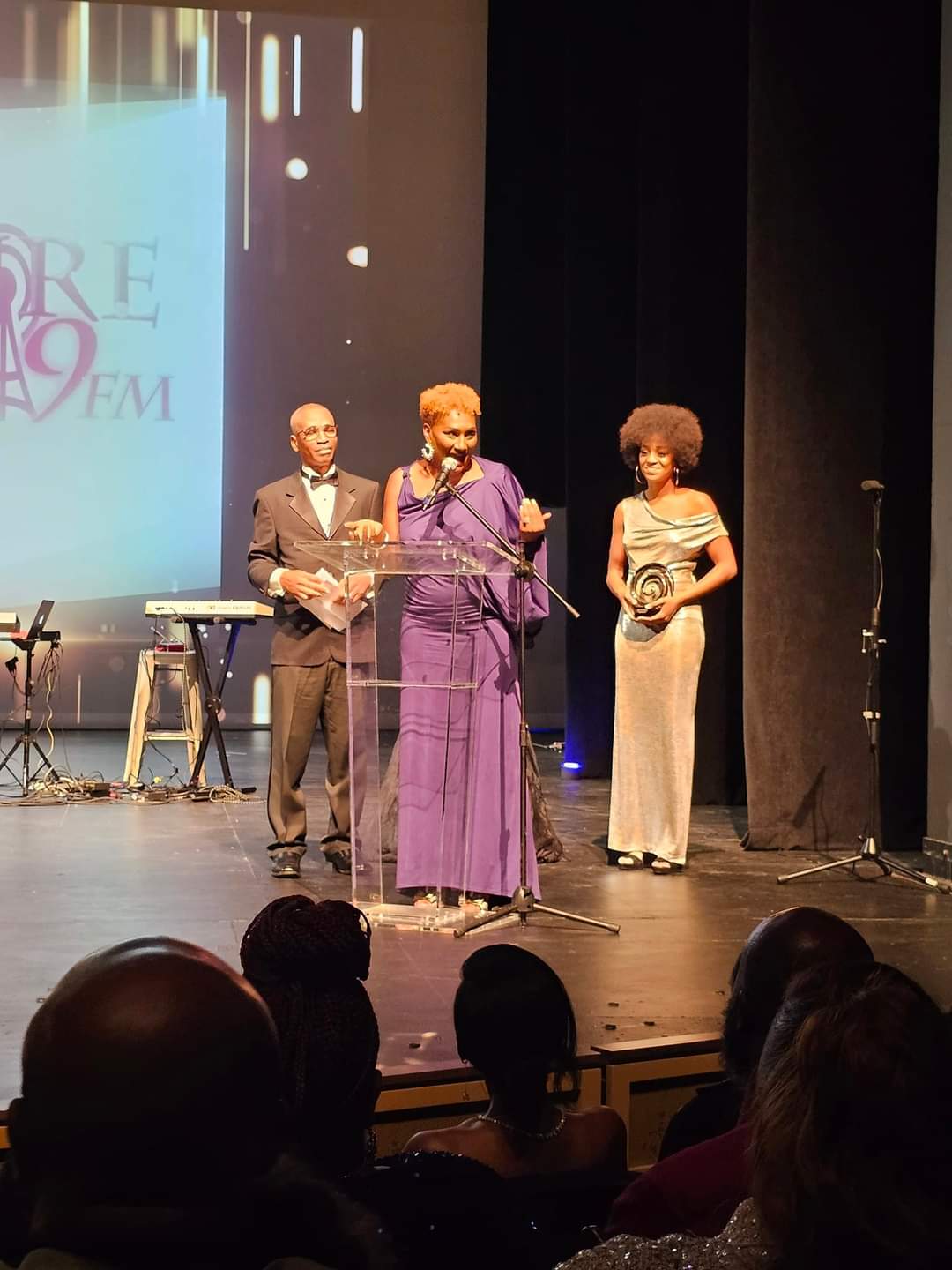 Three people on stage, one speaking at a microphone, two others standing nearby, audience visible in foreground.